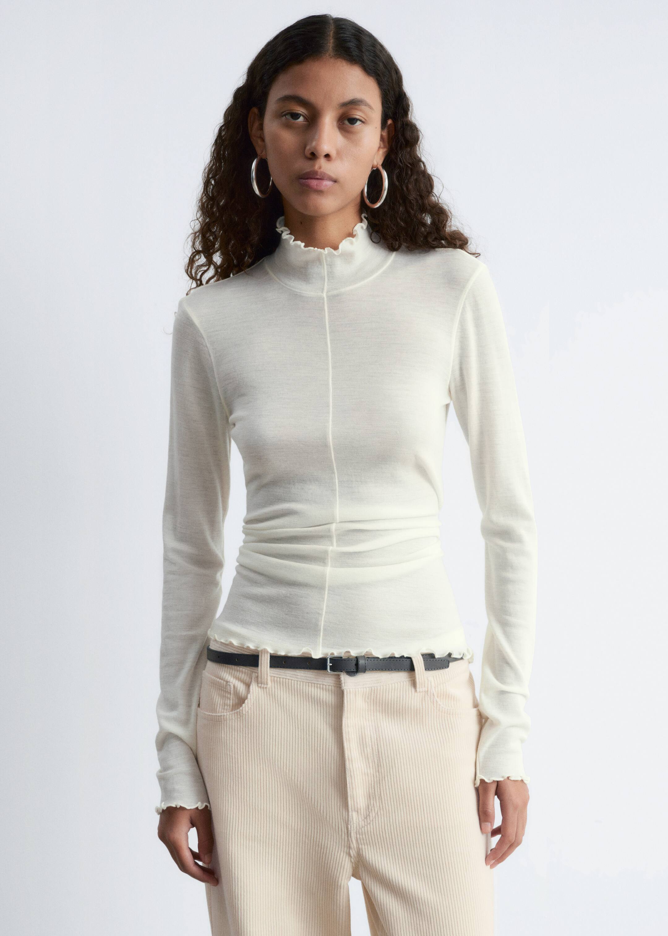 Se større billede: En off-white langærmet top præsenteres forfra med en flæsekrave, flæsemanchetter og en flæsekant. Et synligt lodret søm løber ned langs midten foran, og stoffet fremstår halvgennemsigtigt med en tætsiddende pasform.
