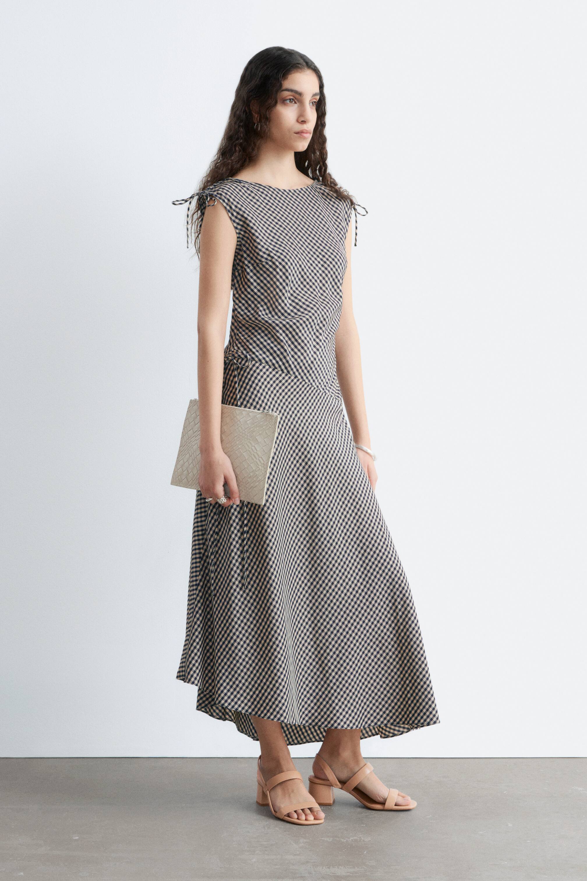 Ingrandisci l'immagine: L'abito presenta un motivo a quadretti scuri e chiari su scala ridotta. È progettato con uno scollo rotondo, dettagli con fiocchi sulle spalle e si stringe in vita con un cordoncino laterale, fluendo in una gonna maxi a trapezio con orlo asimmetrico.
