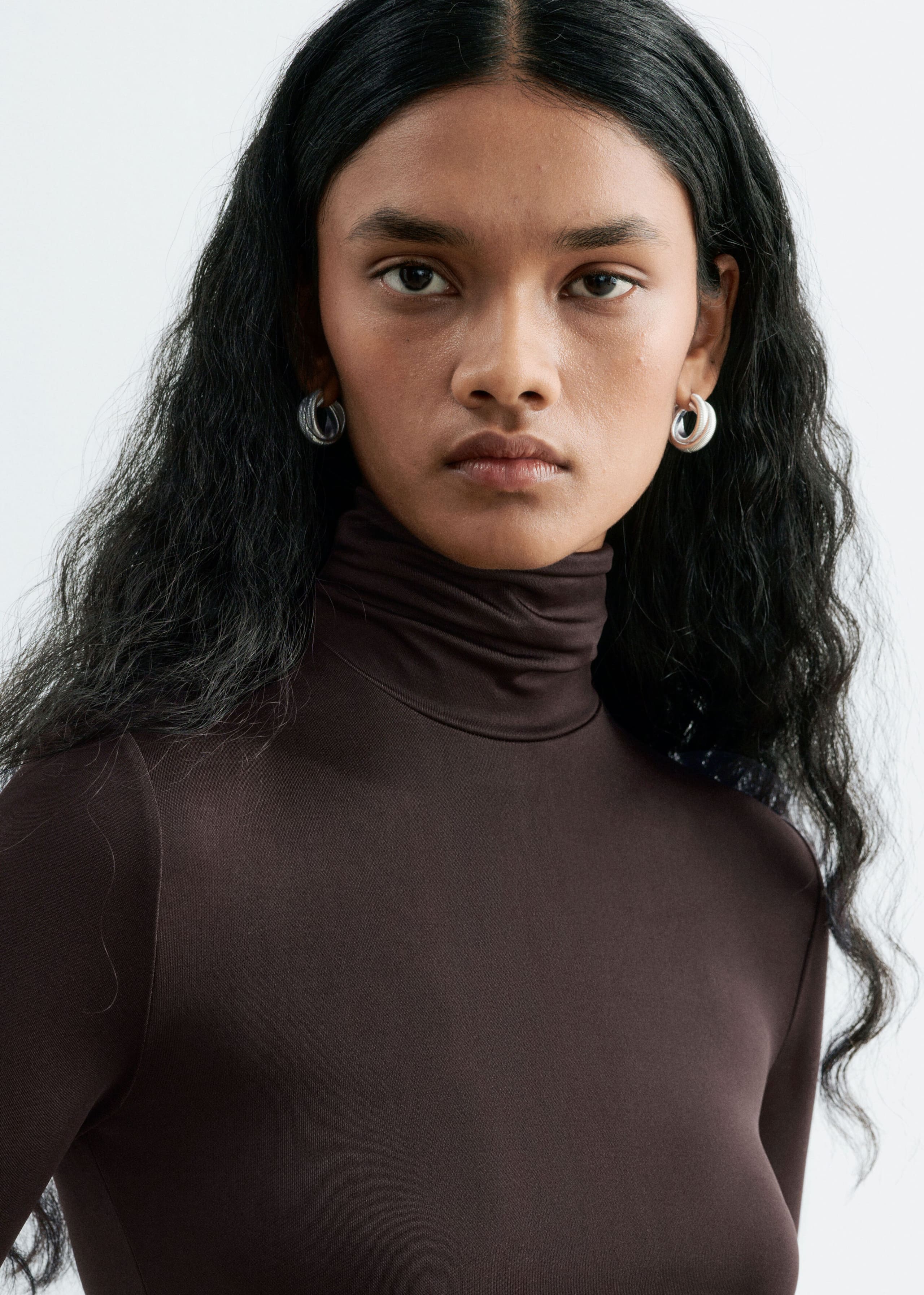 A dark brown jersey dress featuring a double-layered mock neck is being worn by the model in a close-up view. The fitted top of the dress is styled with silver hoop earrings.