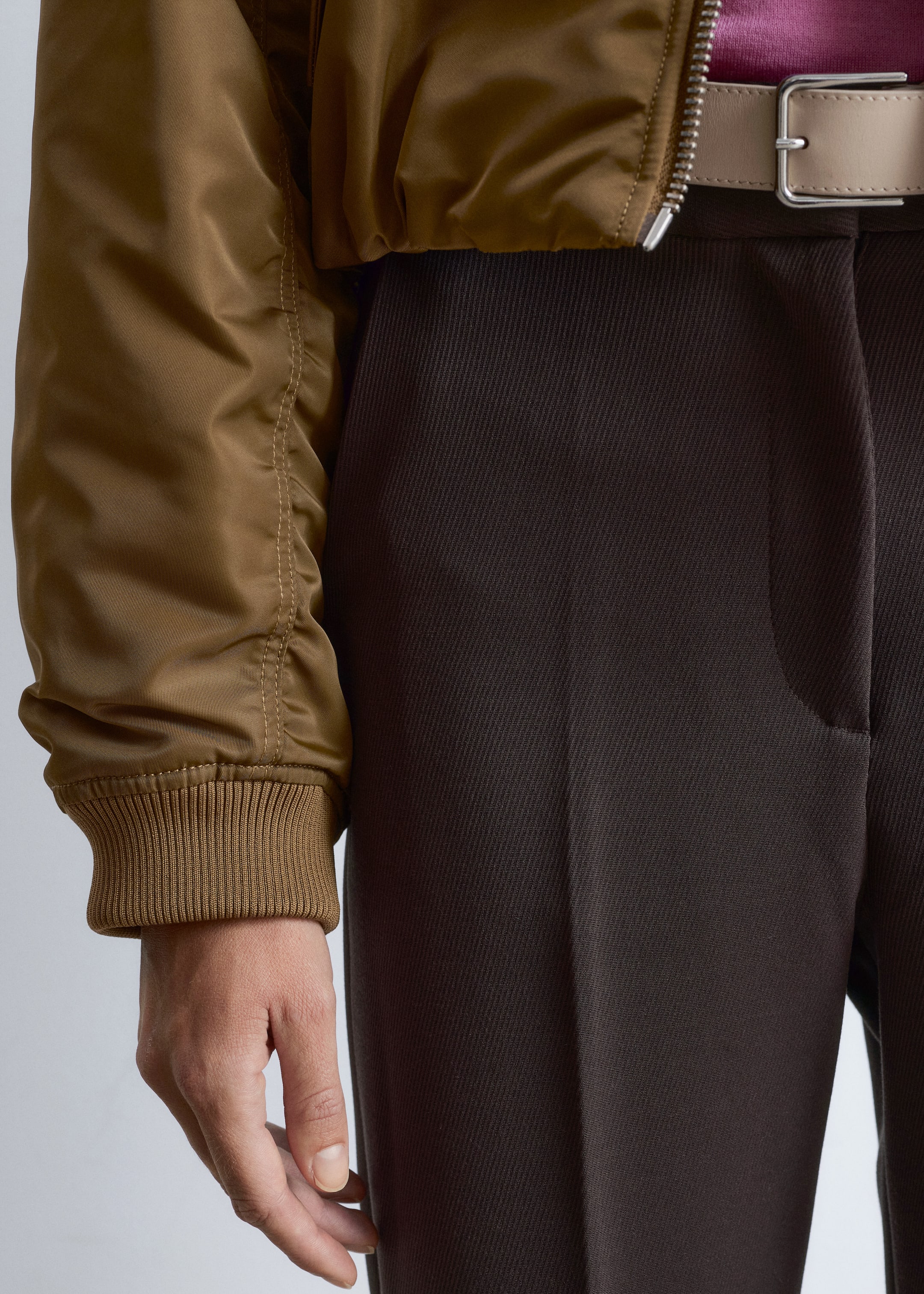 Agrandir l'image: Un pantalon en sergé marron foncé est présenté de côté, montrant sa coupe droite et une ceinture beige clair avec une boucle en argent. Une veste bomber brun olive est superposée à un haut magenta, avec sa manche longue semblant être rassemblée par un poignet côtelé au niveau du poignet.