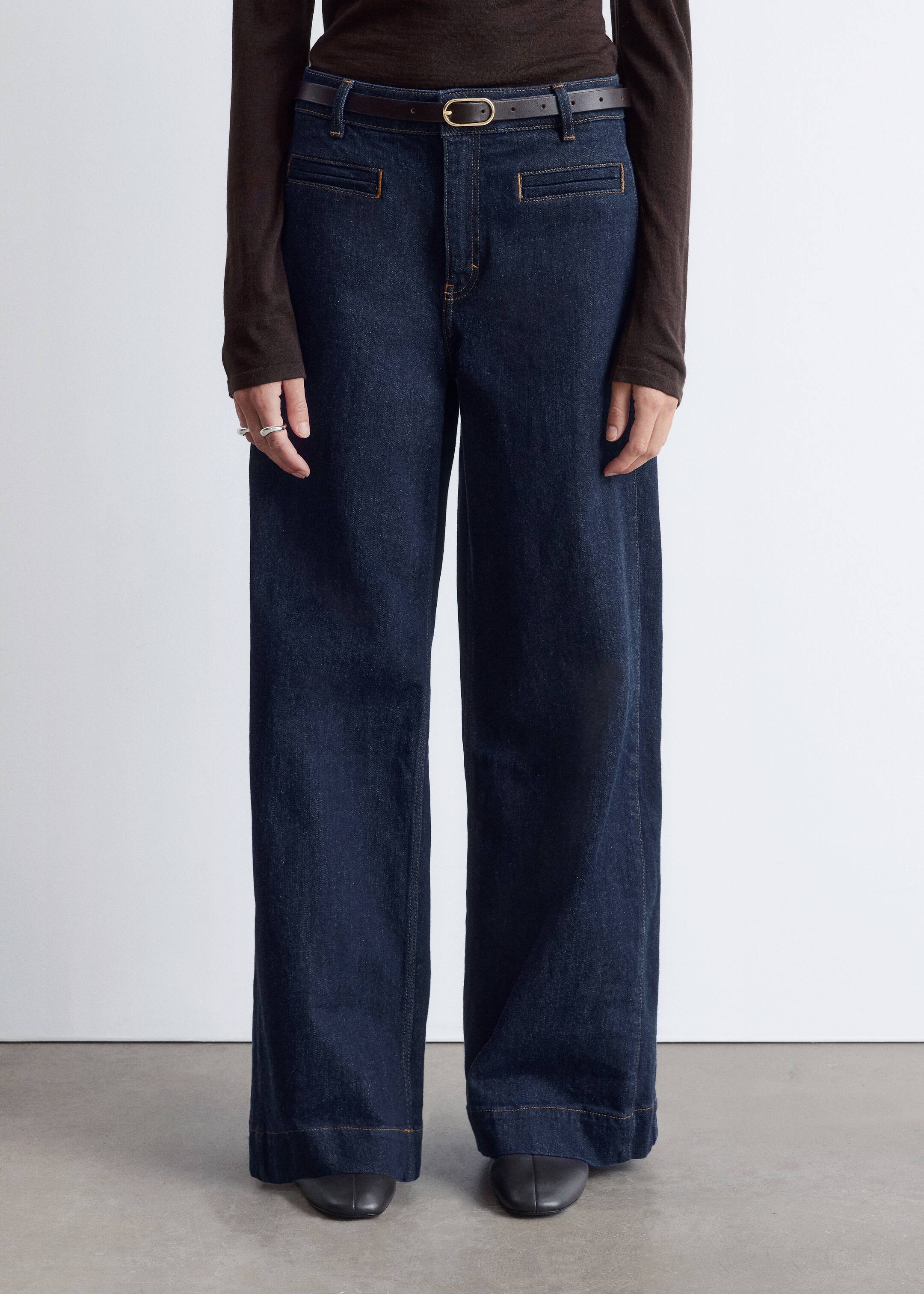 Visa större bild: Mörktvättade jeans med vida ben presenteras från en framåtvänd vinkel, med stiliserade rektangulära framfickor med horisontella öppningar och bärs med ett mörkt läderbälte som har ett ovalt mässingsspänne.