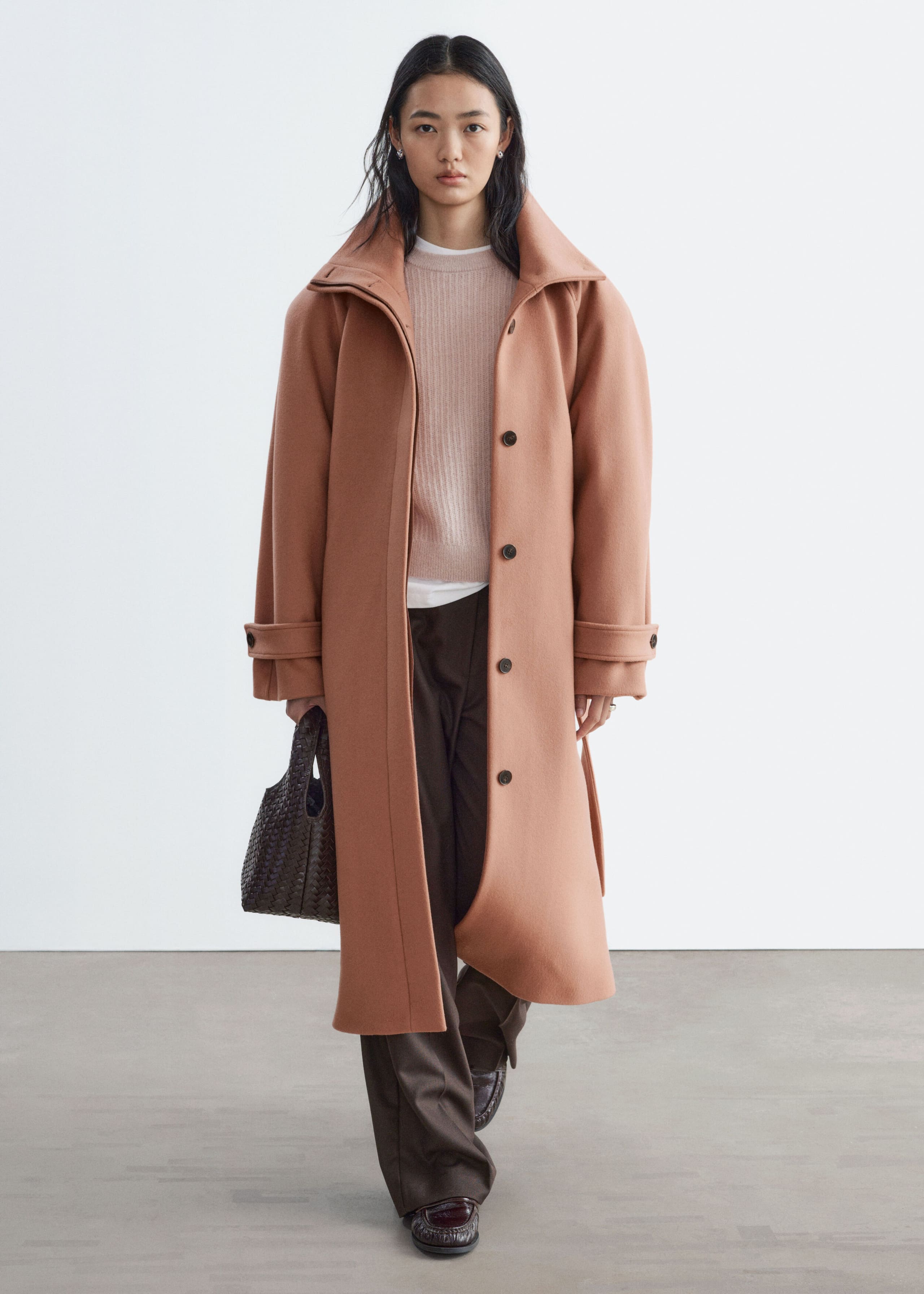 A long, terracotta-pink wool-blend coat featuring structured shoulders, a wide collar and buttoned cuffs is worn open by the model. The coat is layered over a light pink knitted jumper and dark trousers, while the model is holding a dark brown woven handbag.