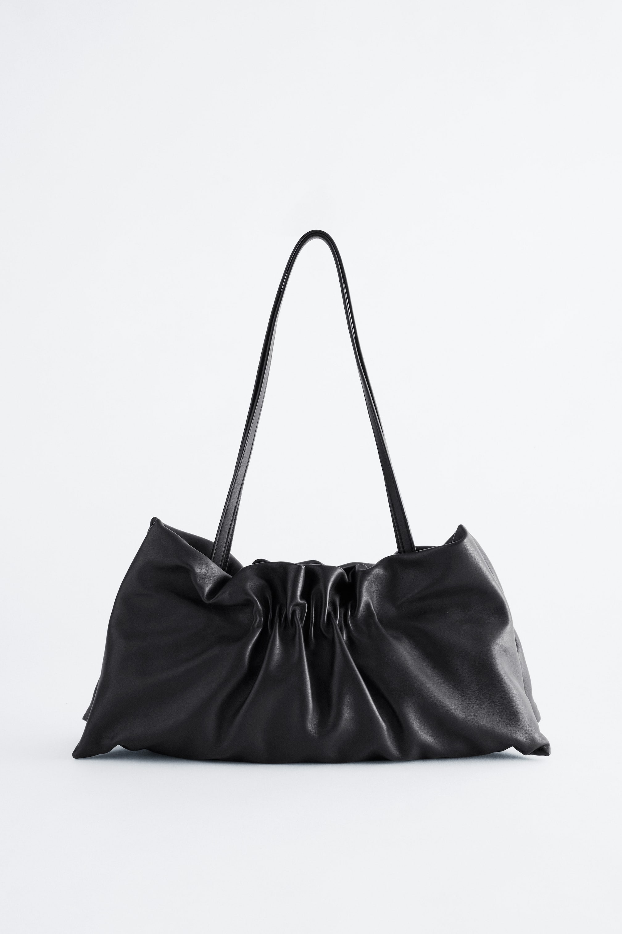 View larger image: A black leather shoulder bag is shown from the front, featuring a soft, slouchy silhouette with ruched, gathered detailing at the centre. Two long, thin shoulder straps arch upwards from the top of the bag.