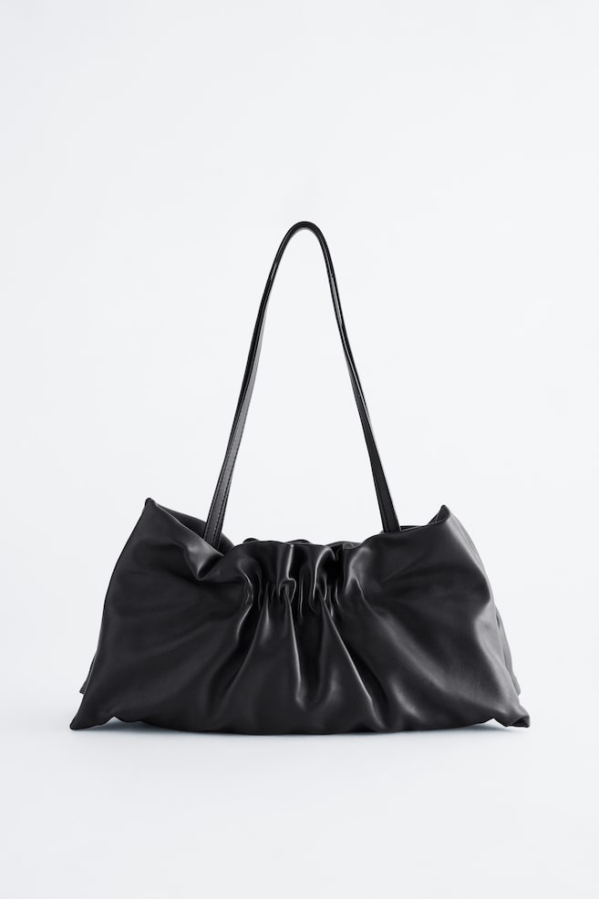 Un sac bandoulière en cuir noir est présenté de face, avec une silhouette souple et décontractée, agrémentée de détails froncés et plissés au centre. Deux longues bandoulières fines remontent en arceau depuis le haut du sac. - 1