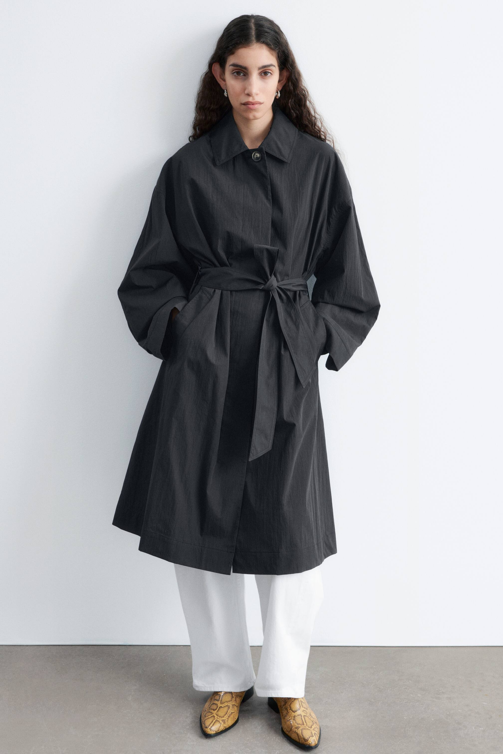 Visualizza immagine più grande: Un cappotto nero presenta un colletto a punta, una chiusura con bottone superiore visibile e una cintura auto-legante che stringe la vita in un nodo. È progettato con maniche ampie e voluminose e tasche laterali dove le mani possono riposare, con il cappotto che arriva fino al ginocchio e mostra una sottile texture verticale.