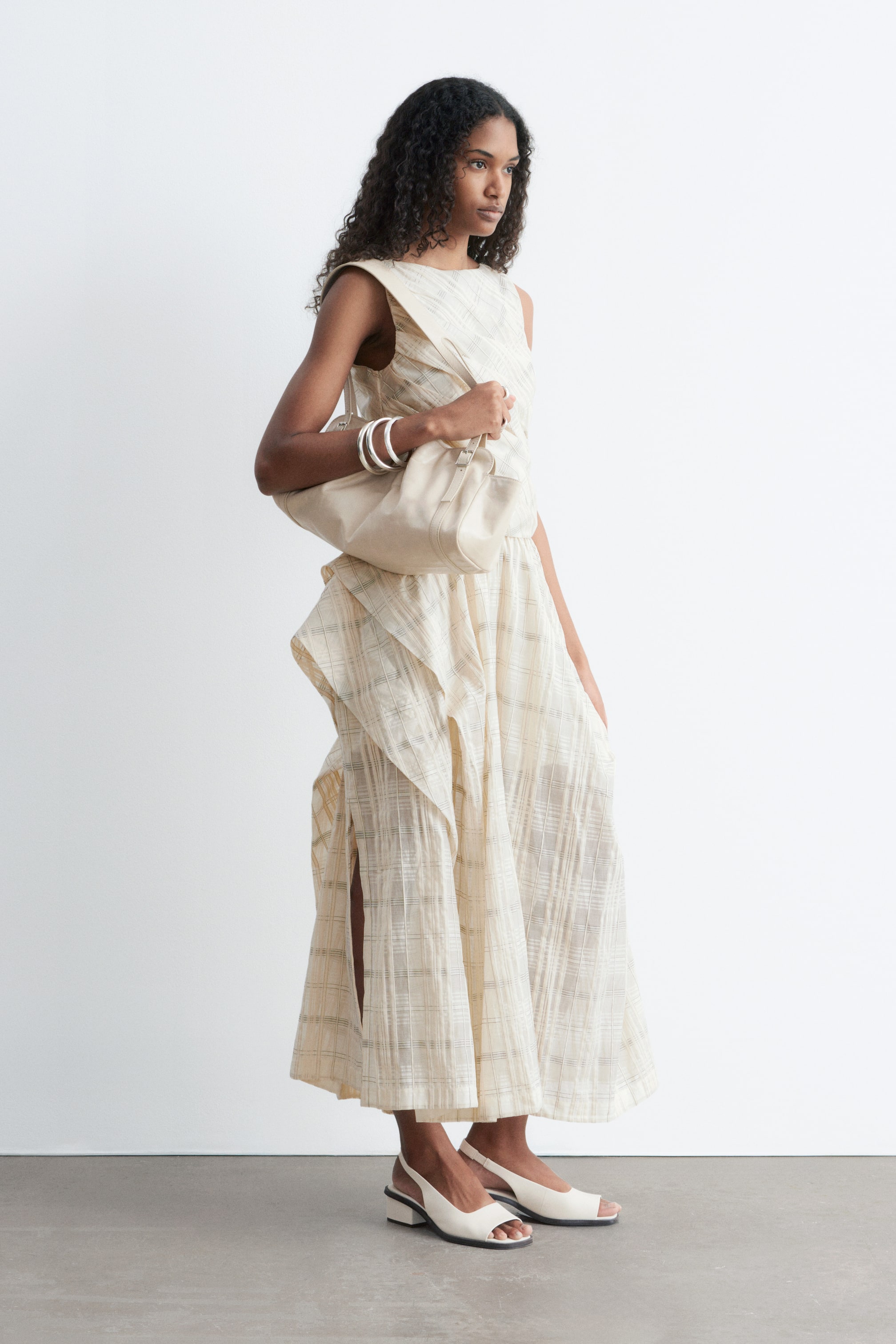 Ver imagen más grande: El vestido es una prenda sin mangas en un color crema claro, que muestra un sutil patrón de cuadrícula. Presenta un escote redondo, un corpiño ajustado y una falda fluida y texturizada con una abertura alta en el lateral, creando una silueta drapeada.