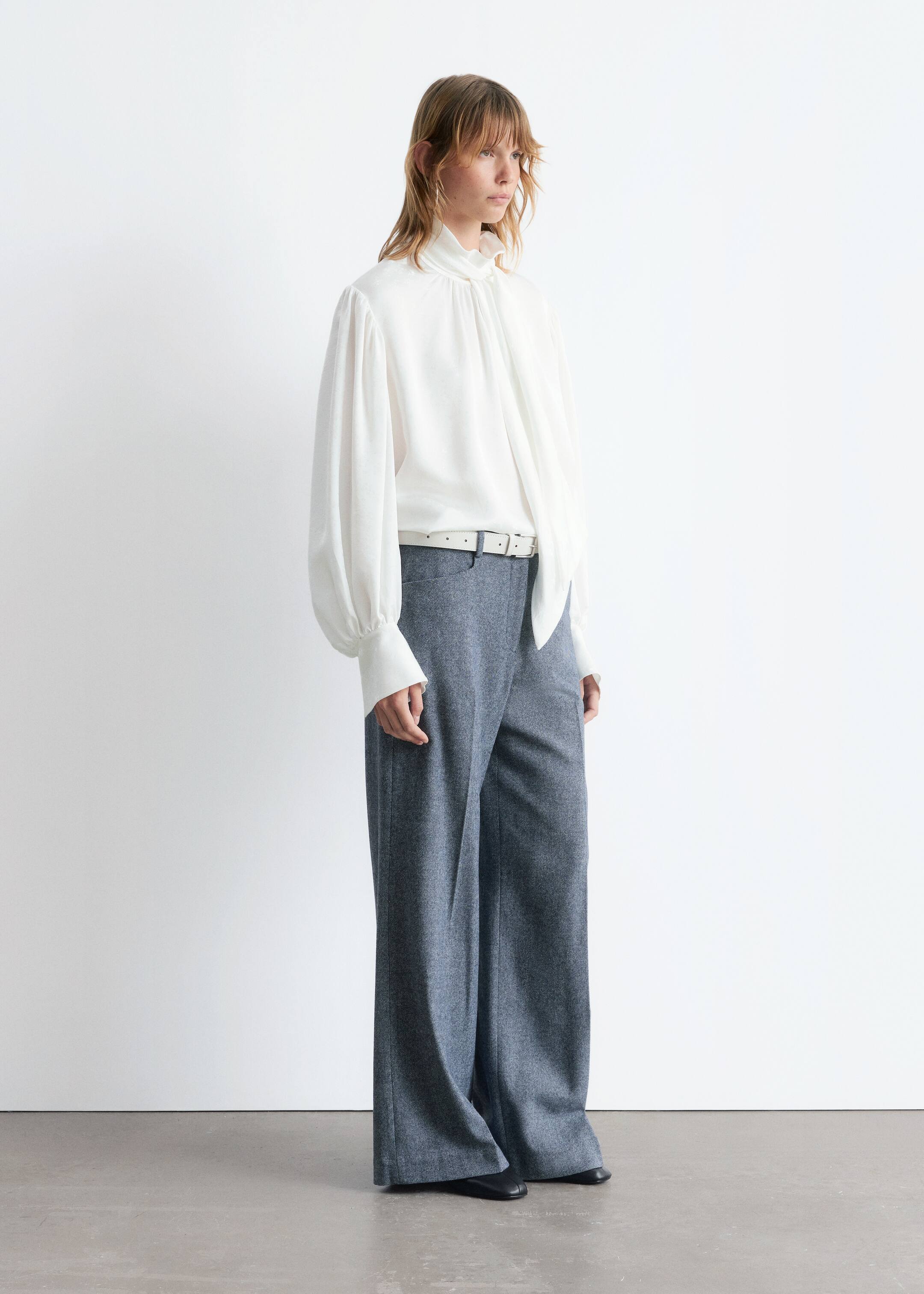Agrandir l'image: La tenue comprend un chemisier blanc avec un col montant à volants, un long détail de cravate tombant sur le devant, et des manches ballon volumineuses avec de longs poignets. Elle est présentée rentrée dans un pantalon gris à jambes larges, avec une ceinture blanche serrant la taille, et des ballerines à bout carré noires portées.