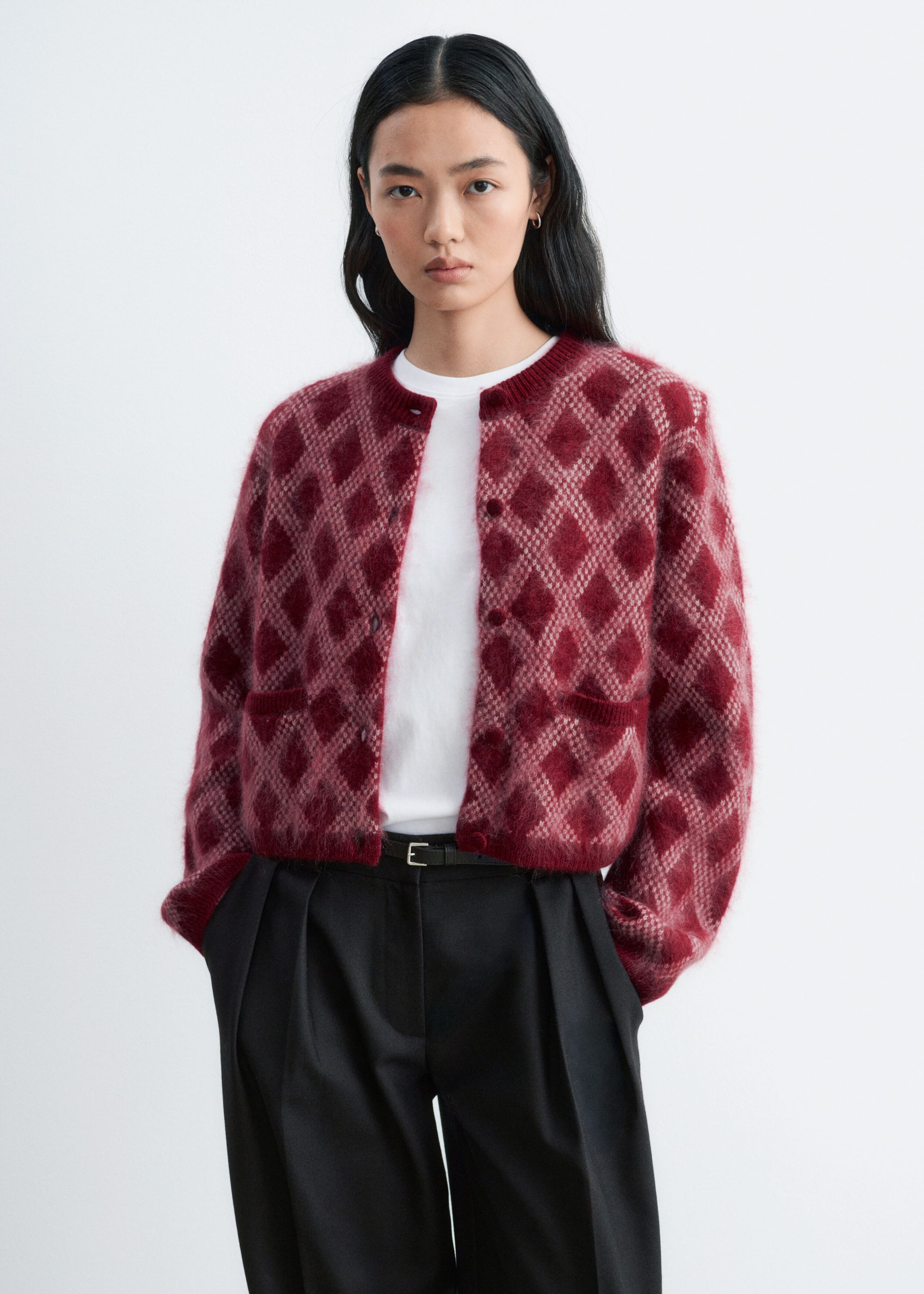 A fuzzy knit cardigan featuring a red and pink diamond pattern is worn open by the model, layered over a white crewneck T-shirt and black trousers. The cardigan has a structured shoulder silhouette, ribbed trims, and small front pockets.