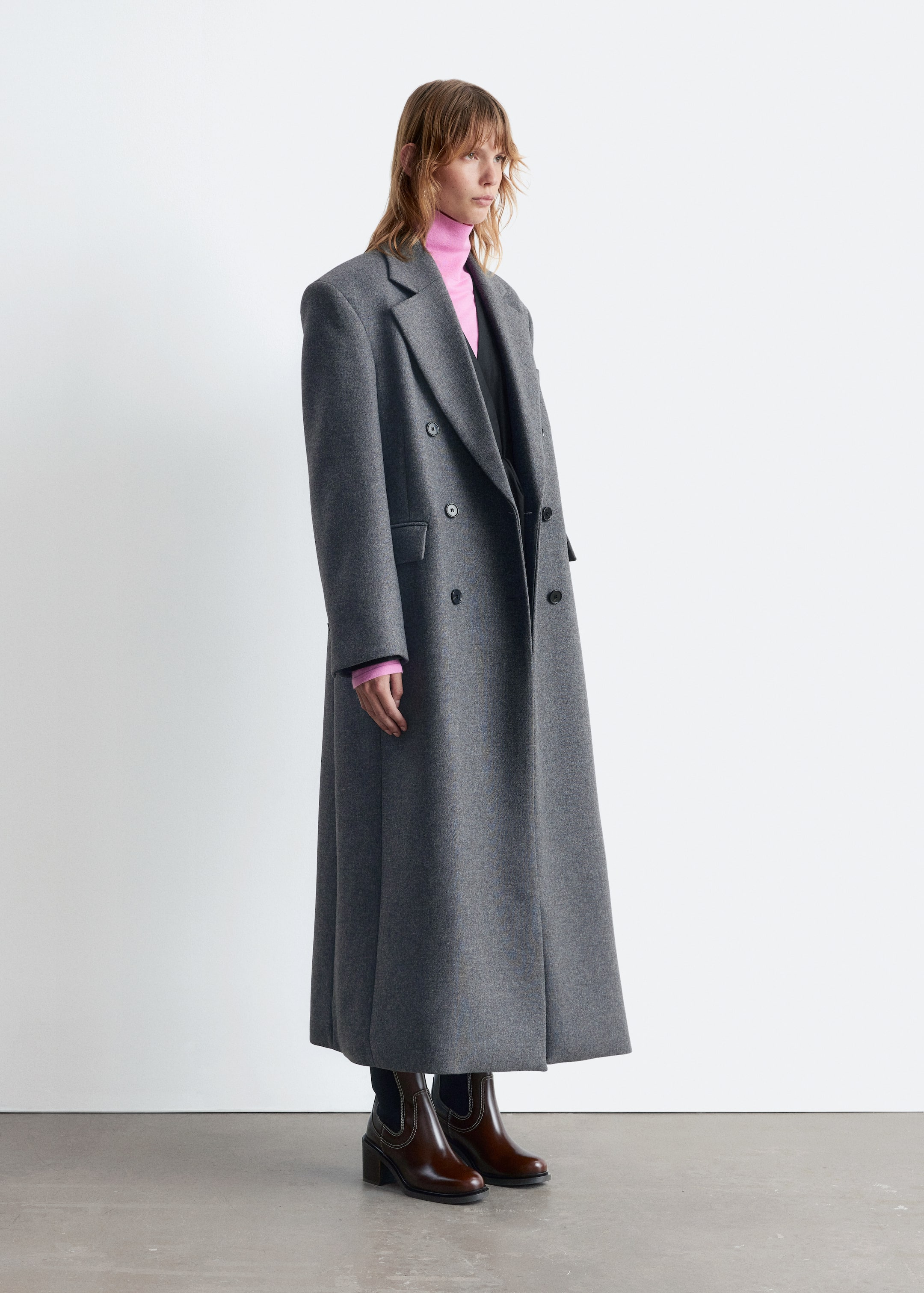 Agrandir l'image: Le manteau maxi gris à double boutonnage est présenté sous un léger angle latéral, avec des revers à encoches larges et proéminents, des épaules structurées et deux rangées de boutons sombres. Il est superposé à un col roulé rose et un vêtement sombre, qui sont visibles en dessous.