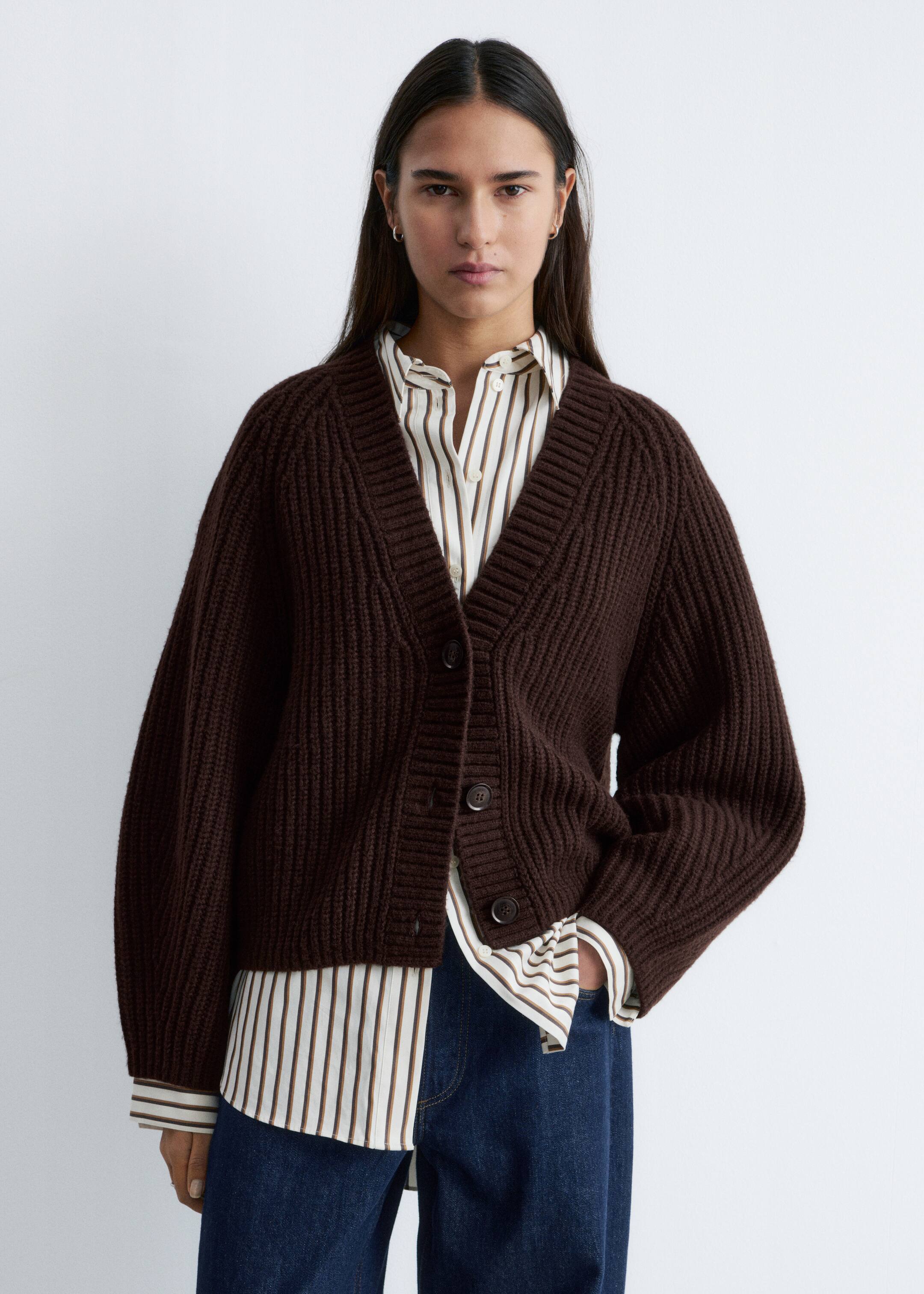 Ver imagen más grande: Un cárdigan grueso de punto acanalado de color marrón oscuro presenta un escote en V y se abrocha por delante con botones marrones a juego. Sus mangas largas y amplias terminan en puños acanalados, y el bajo acanalado descansa sobre una camisa de rayas con cuello.