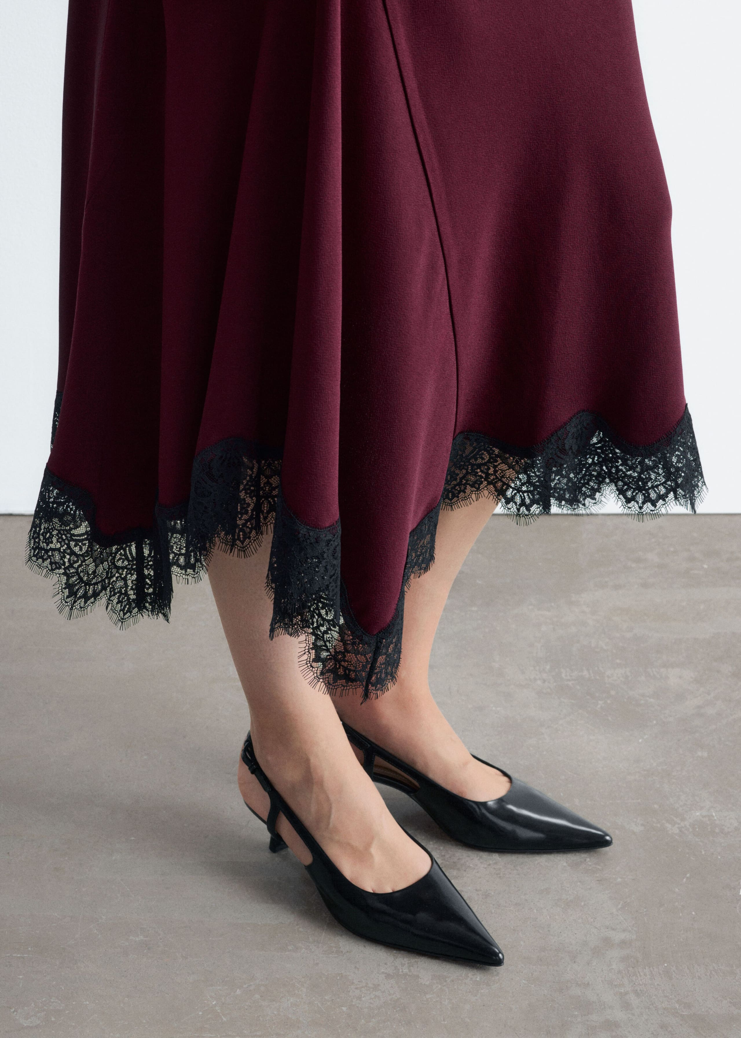 The model is wearing a burgundy flared skirt featuring an asymmetrical hemline trimmed with delicate black lace. The skirt is styled with black pointed-toe slingback heels.