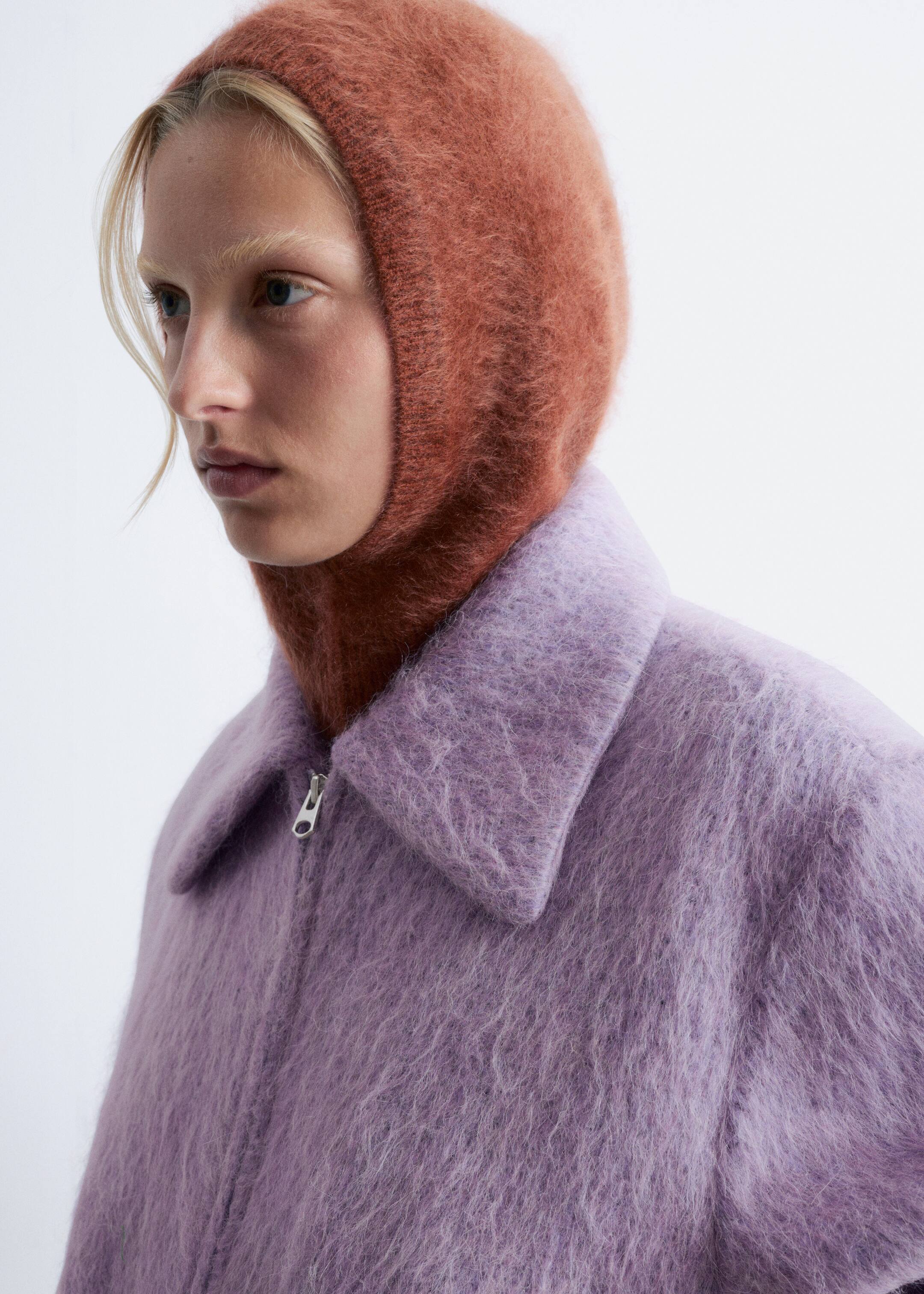 Agrandir l'image: Une cagoule de couleur brun rougeâtre, douce et moelleuse, s'ajuste sur la tête et le cou, mettant en évidence sa texture douce. Elle est portée en couches sous une veste violette claire, qui présente un large col en pointe et une fermeture éclair argentée partiellement dézippée à l'avant.