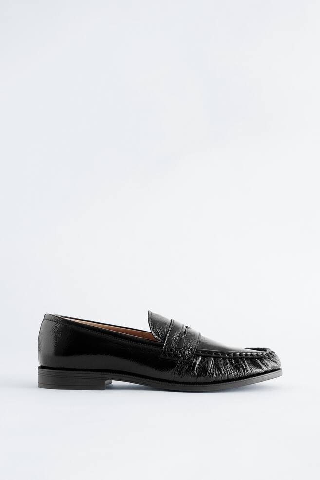 Un mocasín negro de alto brillo se presenta desde una vista lateral, ligeramente inclinado hacia el frente. Presenta una puntera cuadrada con costuras prominentes y una clásica correa de mocasín de penique a través del empeine, y está sostenido por un tacón bajo y plano. - 1
