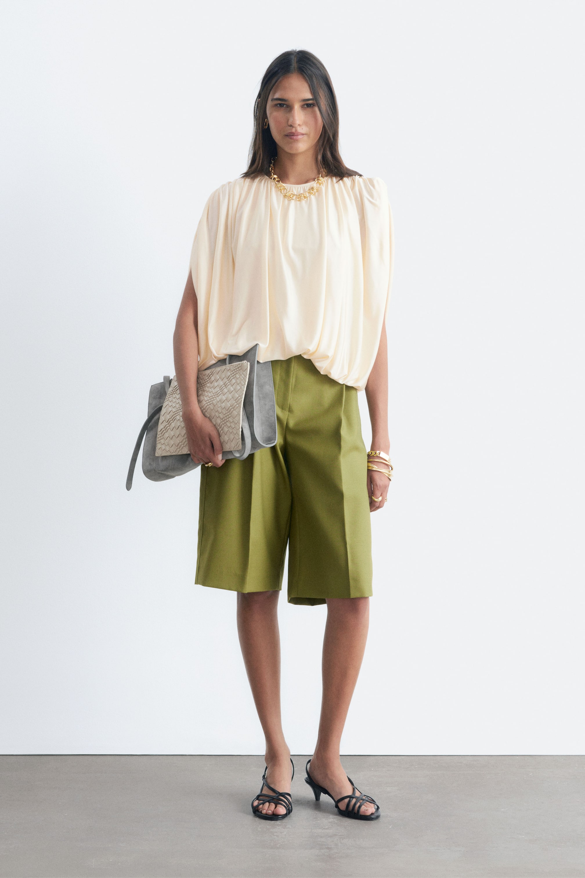 Visualizza l'immagine ingrandita: Una blusa crema drappeggiata con scollo arricciato e maniche ampie è indossata dalla modella. Indossa inoltre bermuda verde oliva, sandali neri con cinturini e gioielli color oro, mentre tiene in mano una pochette intrecciata grigia.
