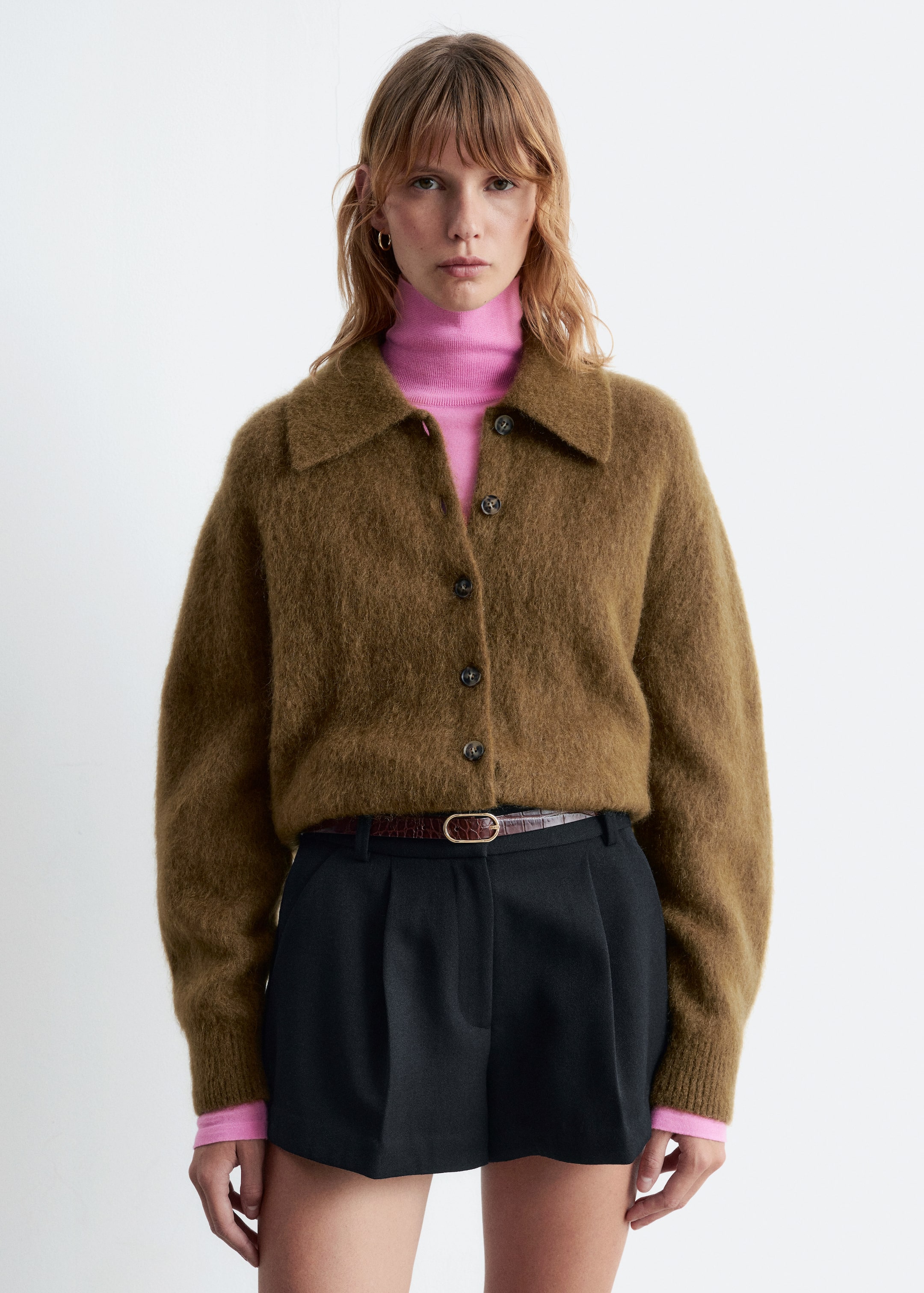 Größeres Bild ansehen: Eine braune, gebürstete Strickjacke wird präsentiert, die einen spitzen Kragen und einen Knopfverschluss mit dunklen, runden Knöpfen aufweist. Sie wird offen über einem rosa Rollkragenpullover getragen, was ihre lockere Passform und die verkürzte Länge betont.