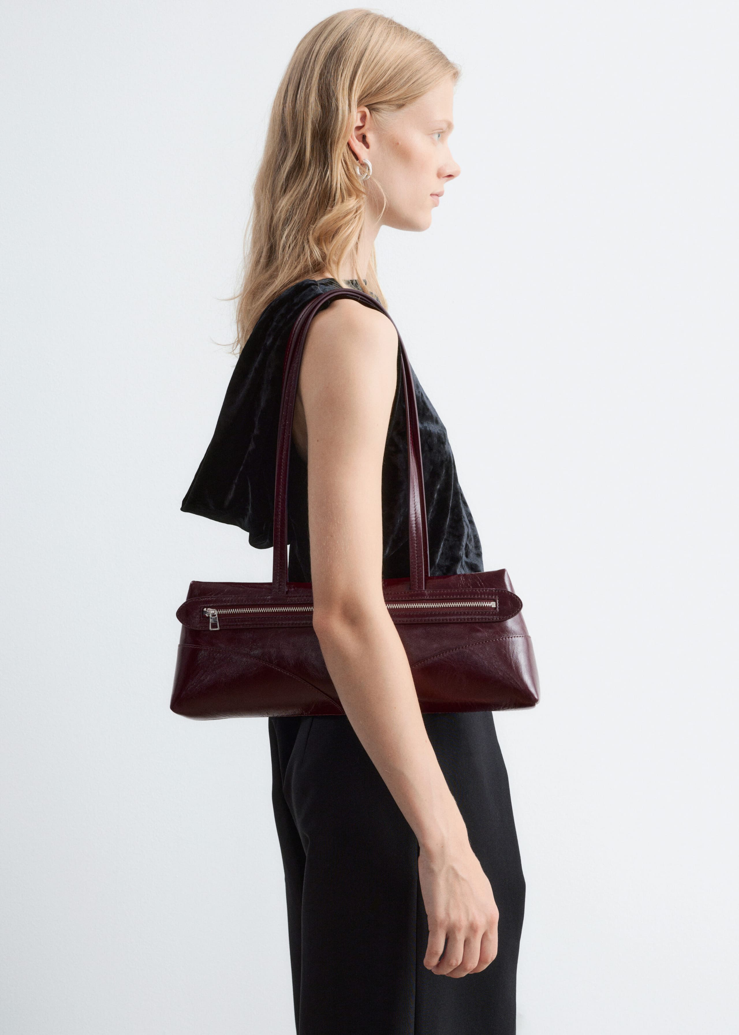A rectangular burgundy leather shoulder bag with a silver front zip fastening and long double straps is being worn over the shoulder by the model. The bag rests at the side, styled with a black sleeveless top and black trousers.