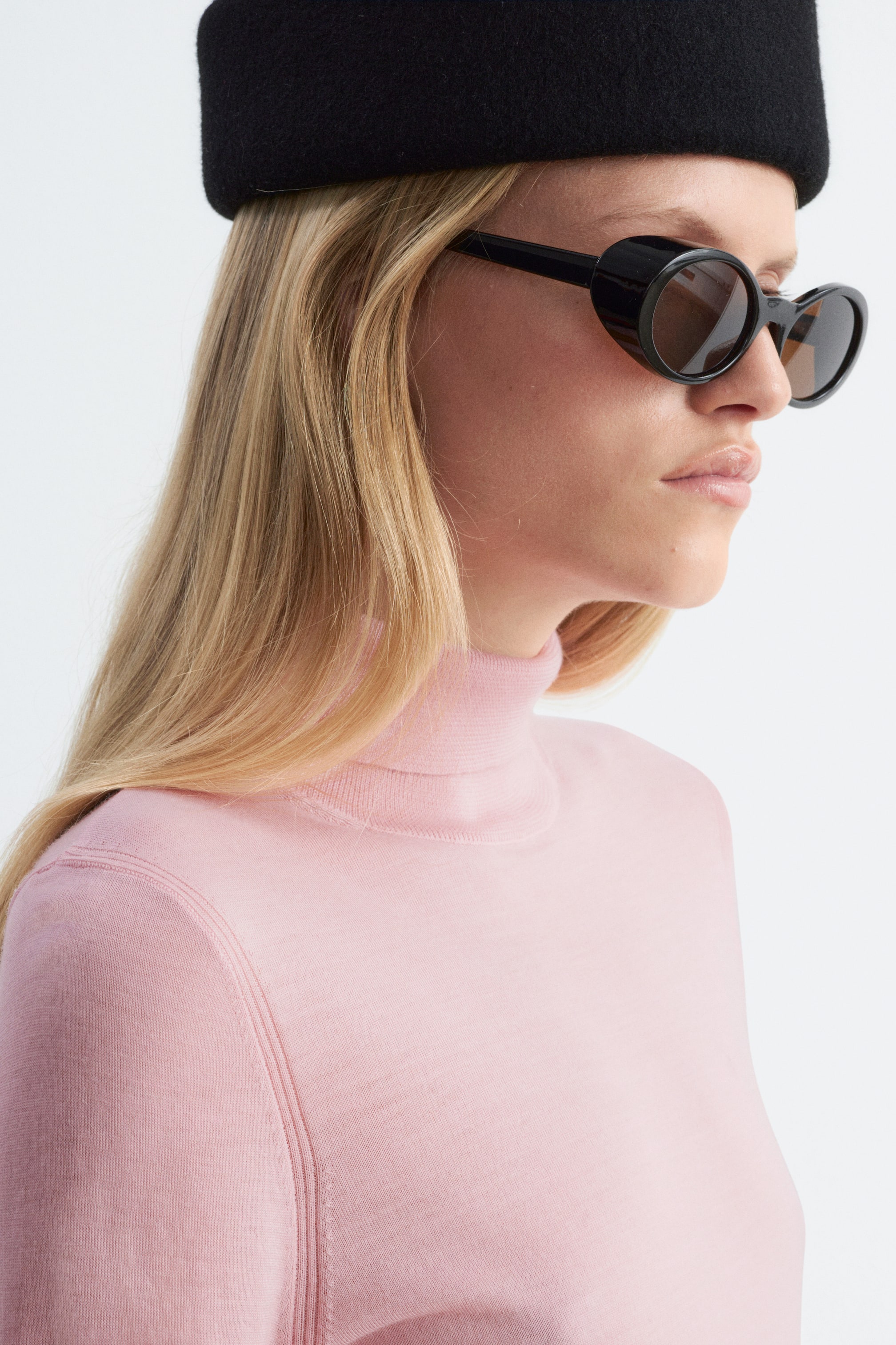 View larger image: The model is wearing a light pink merino wool polo neck top and black oval sunglasses with thick frames. The fine-knit top features a high collar and is styled with a black structured hat.