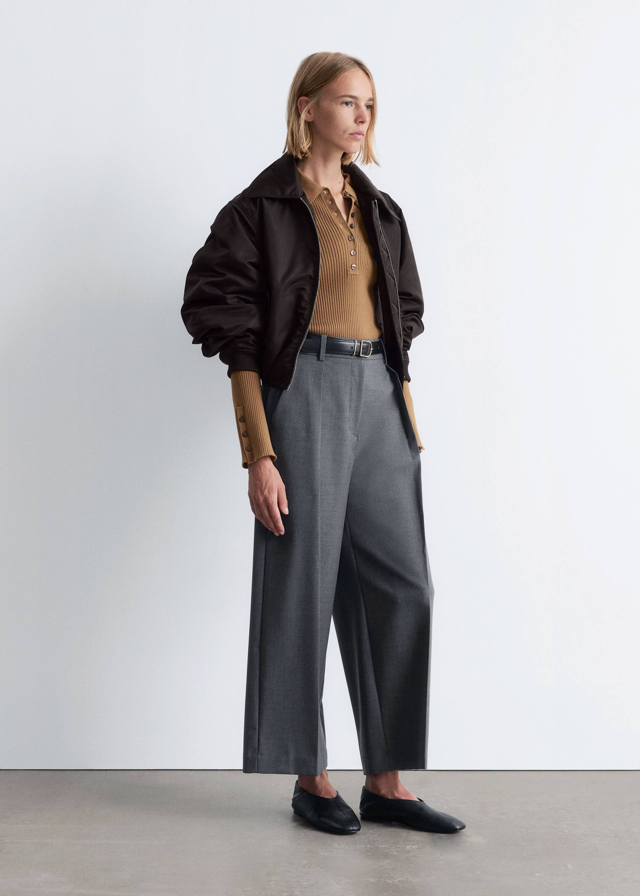 View larger image: A dark brown bomber jacket is worn open over a ribbed tan polo top, displaying a button placket and buttoned cuffs. The top is tucked into grey wide-leg trousers that are cinched with a black belt, and black flat slip-on shoes are completing the ensemble, with the model presenting the outfit from a slight angle.