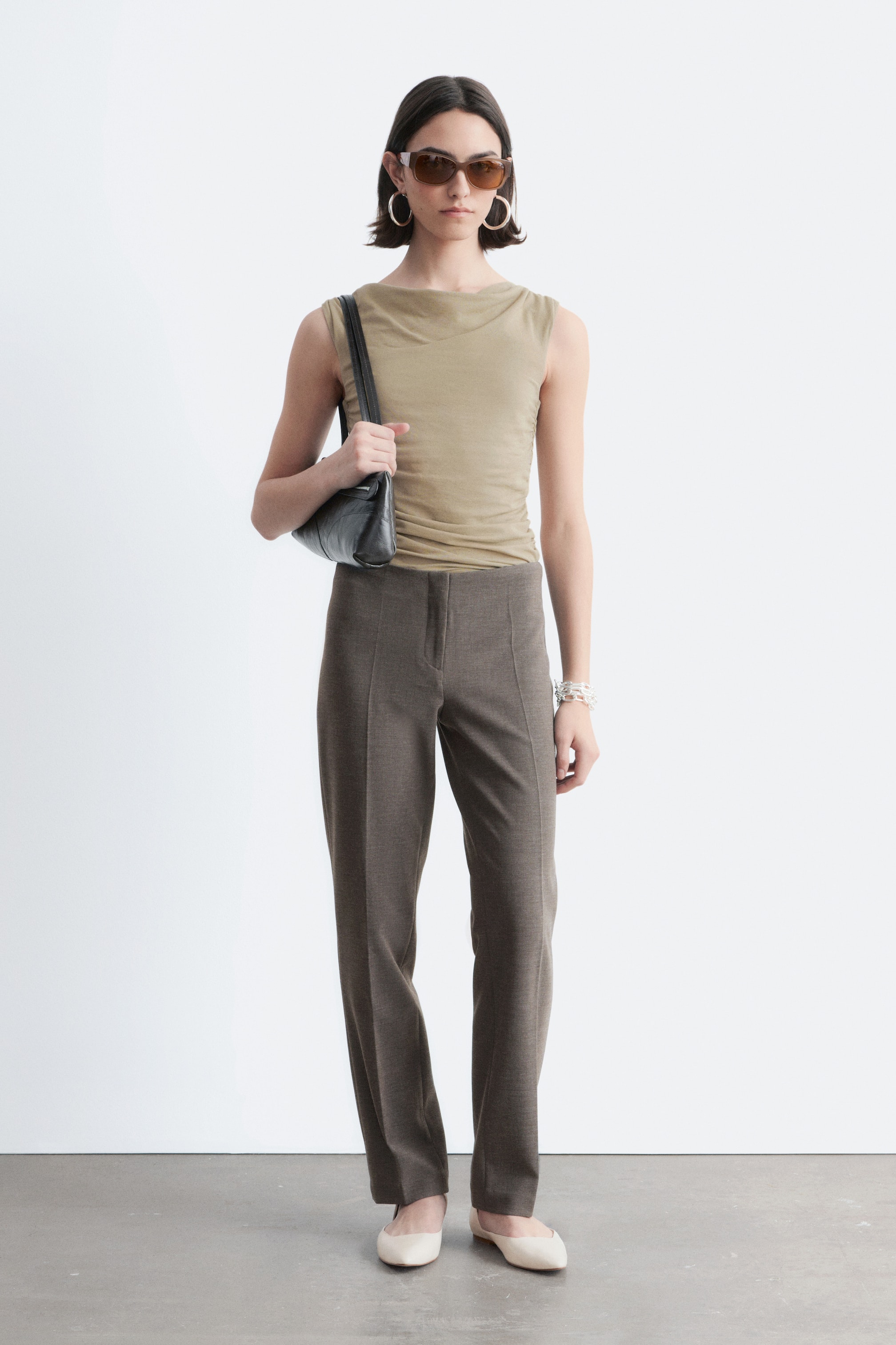 Näytä suurempi kuva: Vaalean oliivinvihreä hihaton toppi, jossa on drapeerattu pääntie ja hienovaraiset rypytykset, esitetään tummanruskeiden suoralahkeisten housujen kanssa, joissa on etusaumayksityiskohta. Vaalean kermanväriset teräväkärkiset balettikengät ja musta tikattu olkalaukku täydentävät kokonaisuuden.