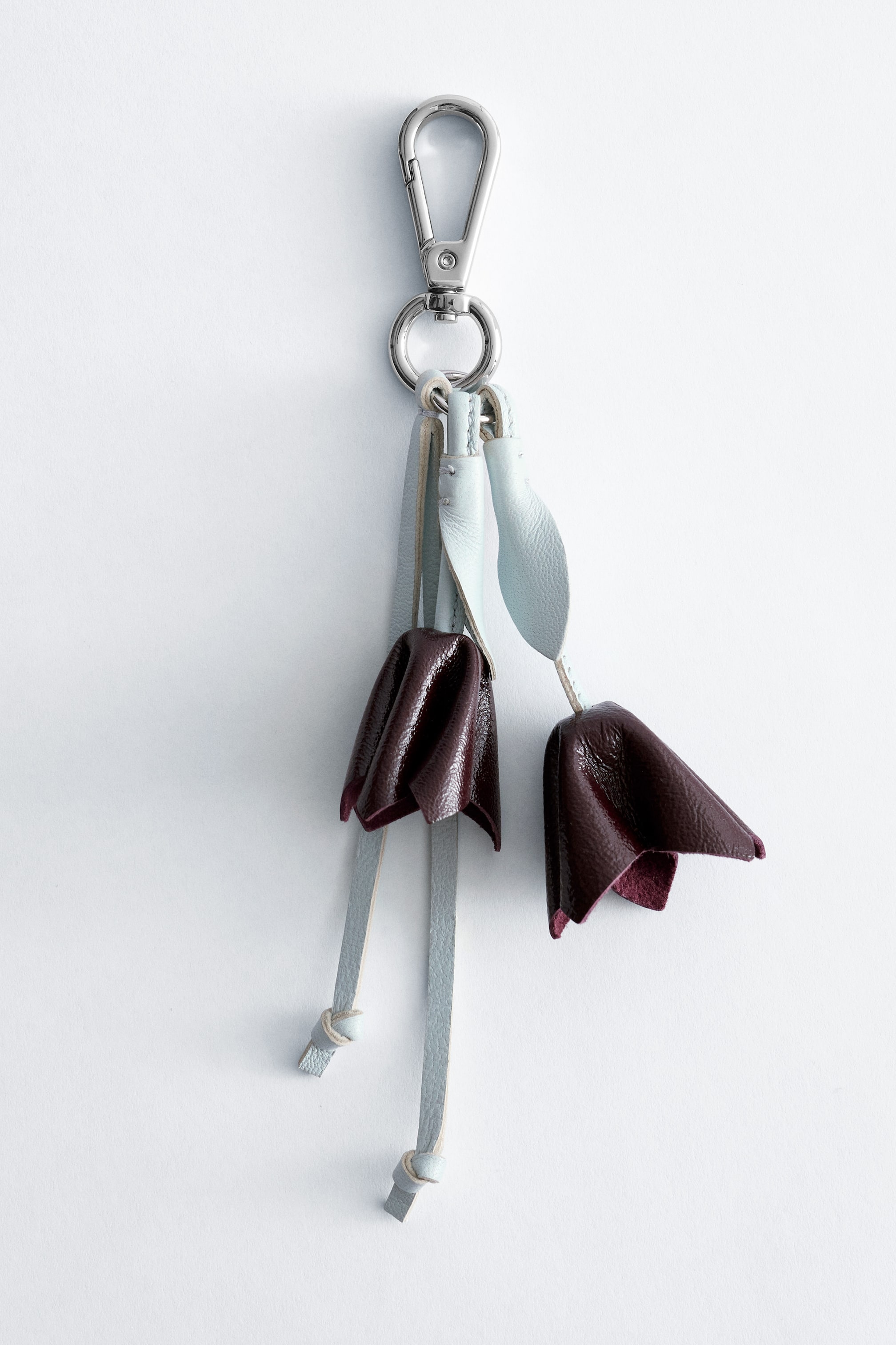 Agrandir l'image: Un charm de sac présente son devant, avec un mousqueton et un anneau de couleur argentée à son sommet, d'où pendent plusieurs lanières en cuir bleu clair. Deux décorations en cuir rouge foncé, froissé et en forme de tulipe, sont suspendues à deux de ces lanières, et d'autres lanières en cuir bleu clair avec des extrémités nouées pendent en dessous.