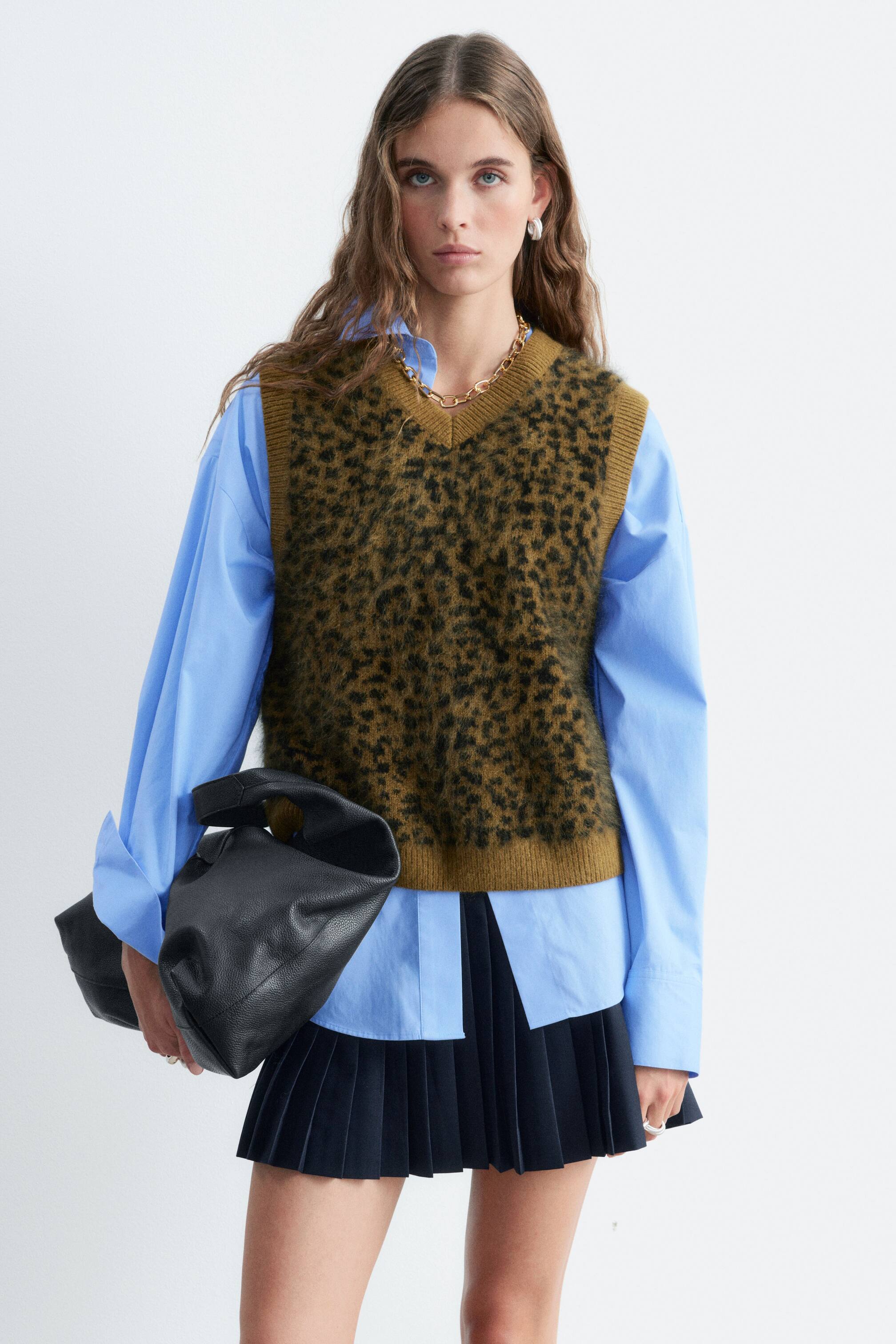 Vezi imaginea mai mare: Un pulover fără mâneci din lână, cu un corp jacard pufos, cu imprimeu leopard, și borduri striate verde-oliv este purtat de model. Puloverul fără mâneci cu decolteu în V este purtat peste o cămașă bleu cu nasturi și stilizat cu o fustă închisă la culoare, plisată.