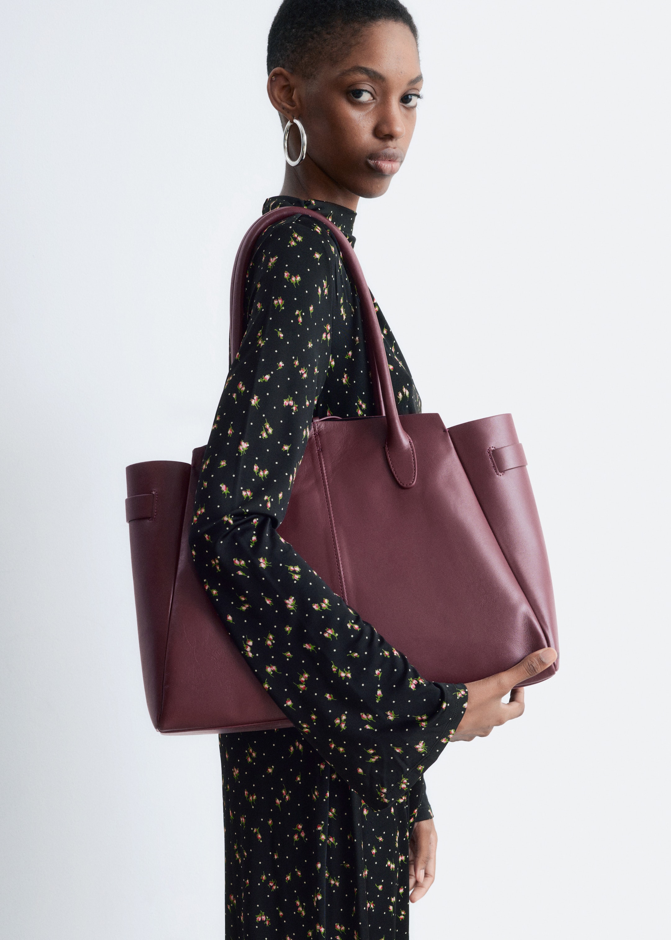 View larger image: A burgundy tote bag is being held by its top handles, facing slightly to the front and side. It is featuring a smooth texture and structured sides with decorative tabs.