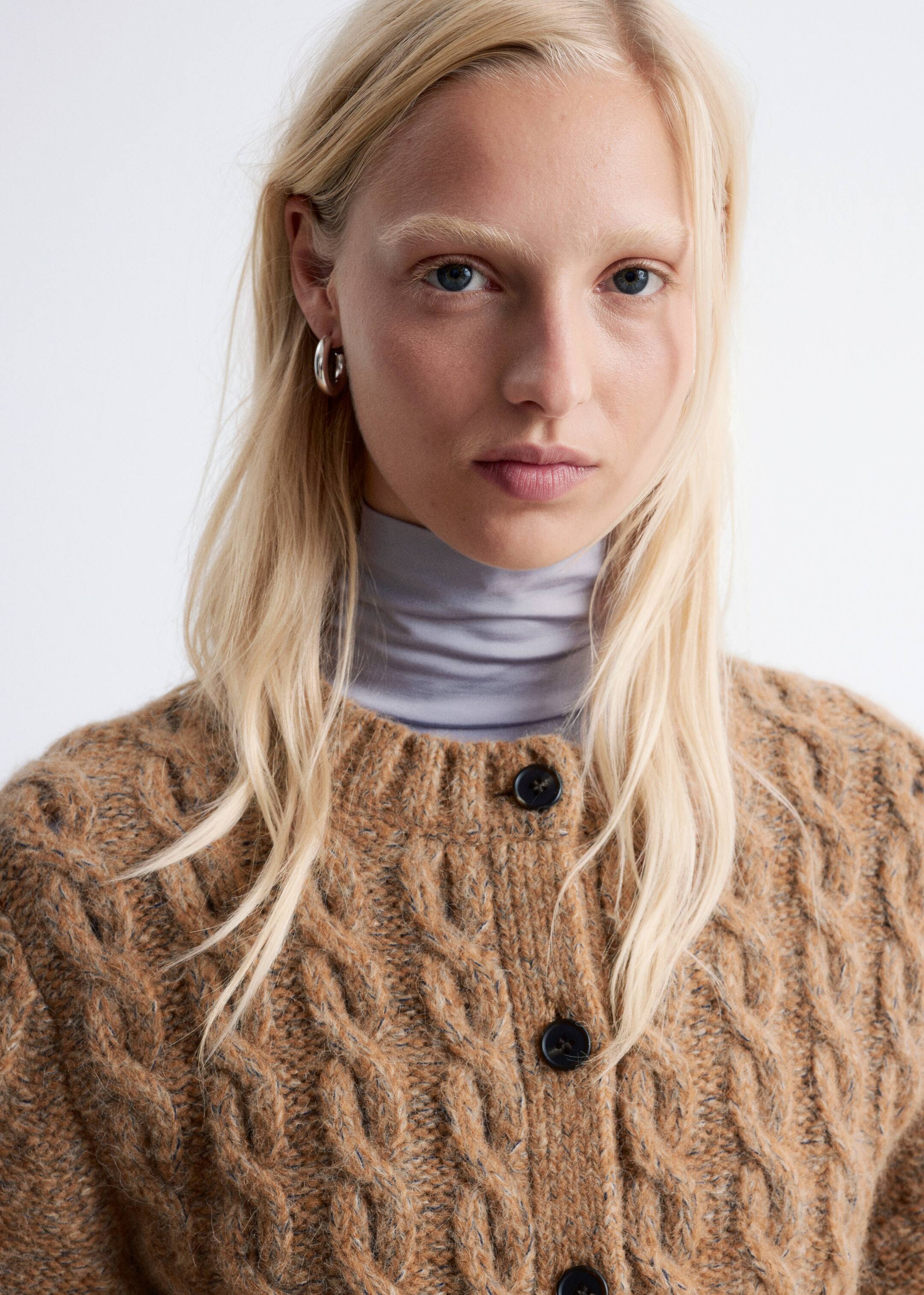 Agrandir l'image: Un cardigan marron à torsades, avec un col rond et des boutons noirs sur le devant, est porté par le mannequin. Le cardigan est superposé à un col roulé gris clair.