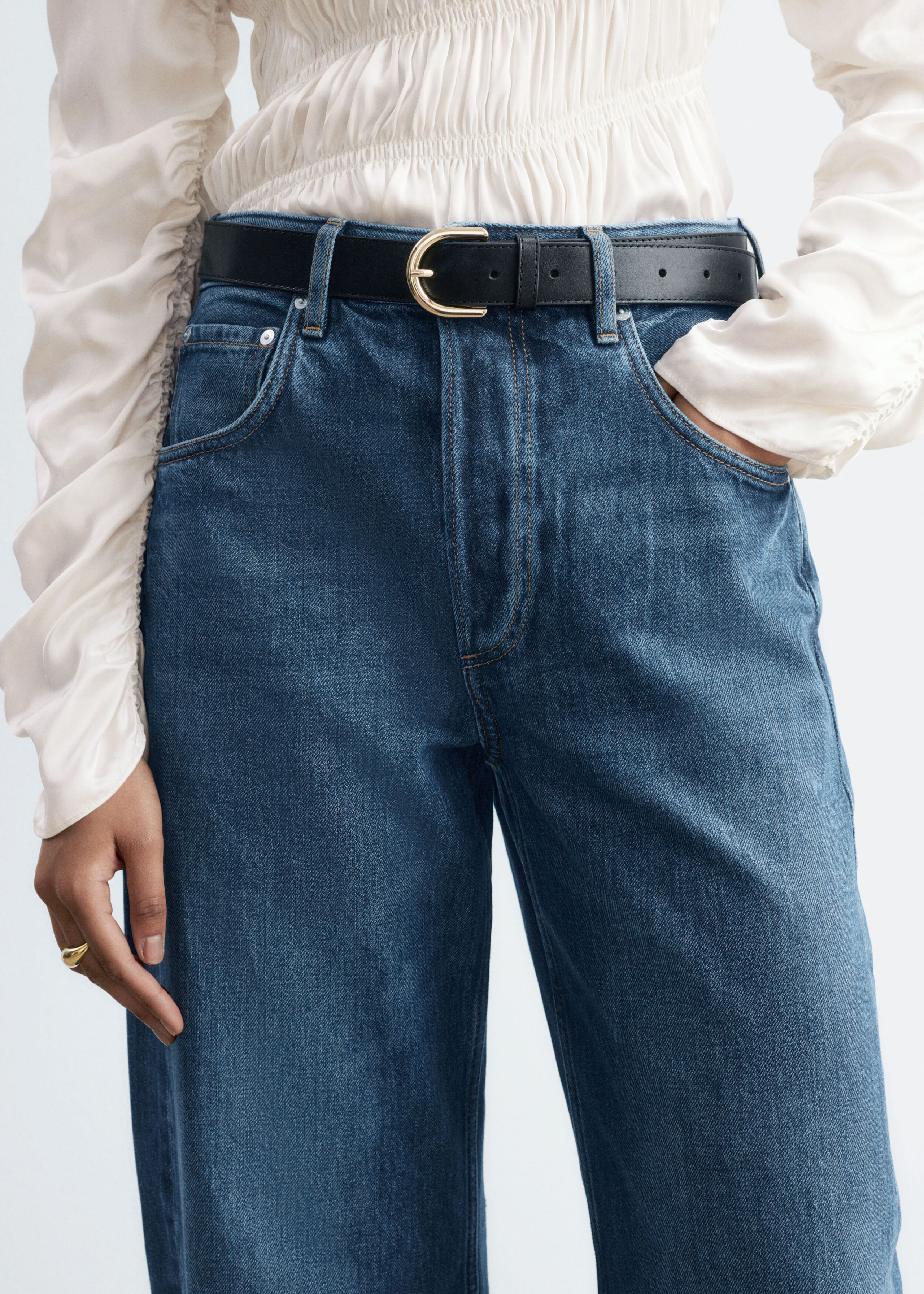 Blue denim jeans featuring a five-pocket design and contrast stitching are worn by the model with a tucked-in cream-colored long-sleeve top and a black leather belt. The model is standing with one hand resting in the front pocket of the mid-rise jeans.