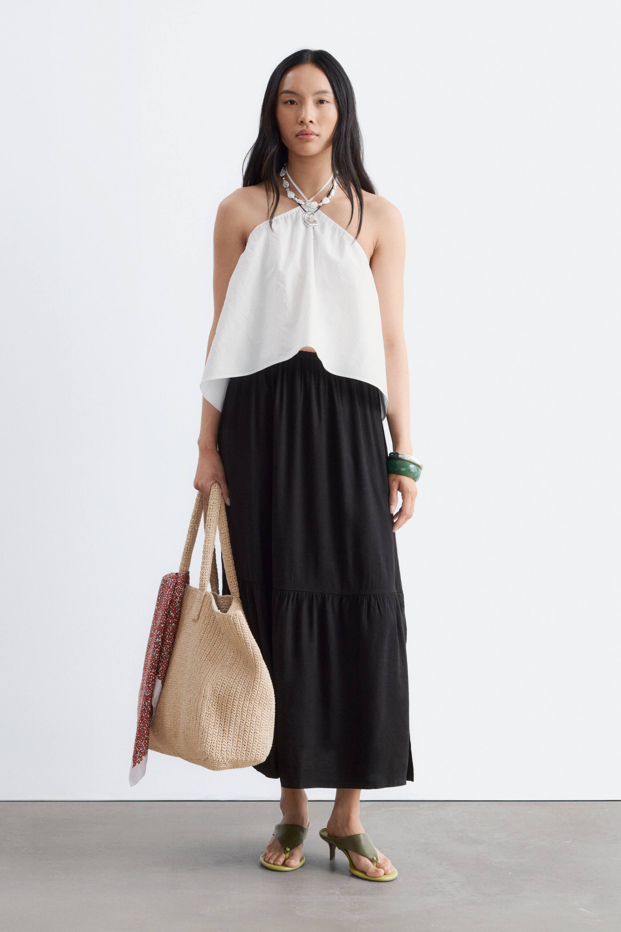 View larger image: A long black tiered skirt is showcased from the front, featuring a relaxed, flowing fit and a gathered waistline. A horizontal tiered seam is visible around the knee, and a subtle side slit appears on the lower left.
