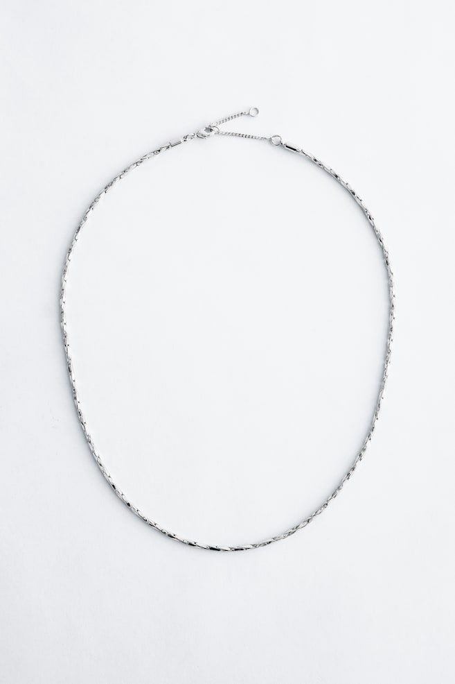 A silver-coloured necklace is lying in an oval shape, featuring a serpentine chain with tightly linked flat, elongated segments. A lobster claw clasp and an extender chain are visible at the top. - 1