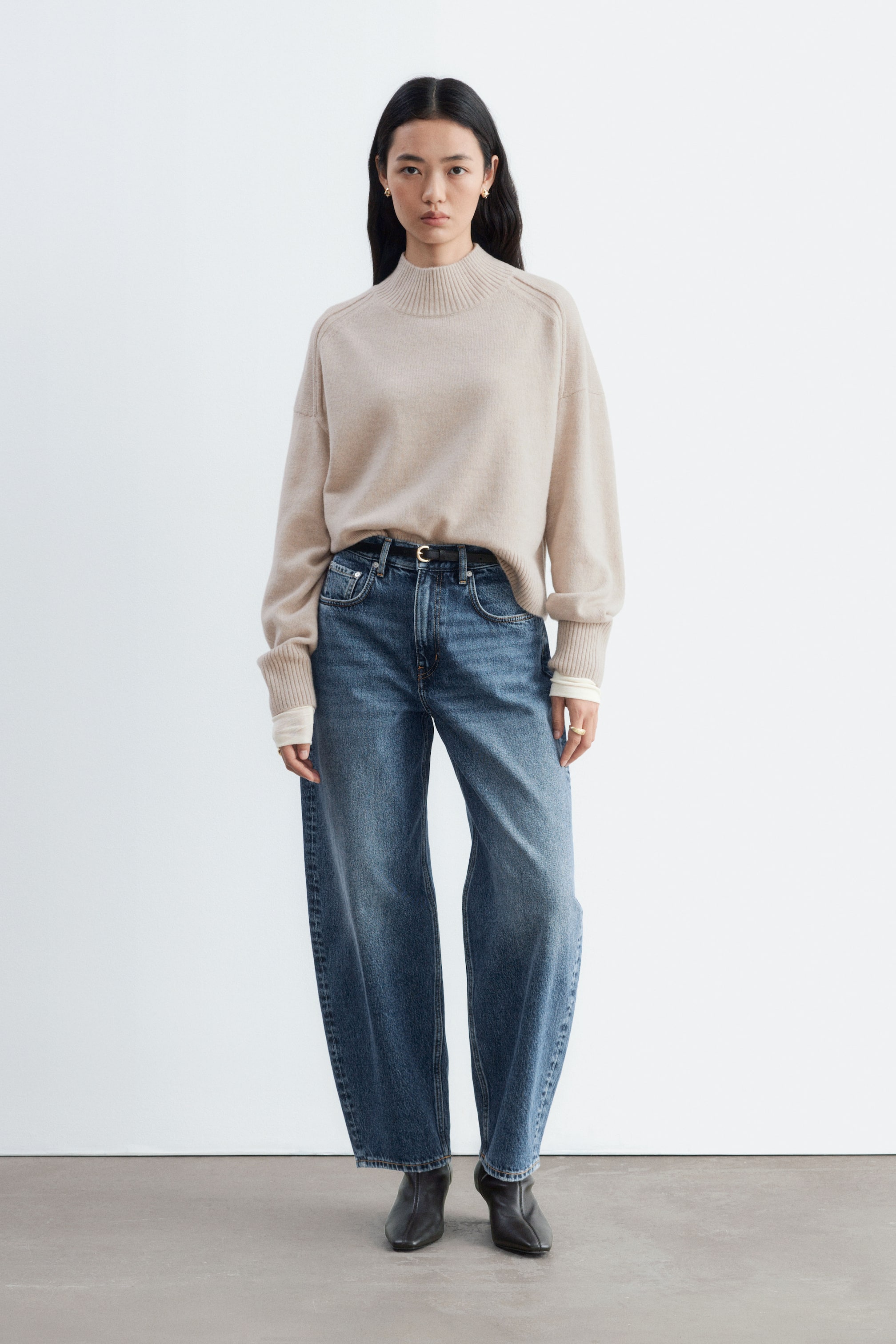 Visa större bild: Barrel-leg ankeljeans i en mellanblå tvätt bärs av modellen. Jeansen med hög midja har ett böjt, avslappnat ben som smalnar av vid ankeln och stylas med en instoppad beige polotröja med falsk hals, ett smalt svart bälte och svarta stövlar.