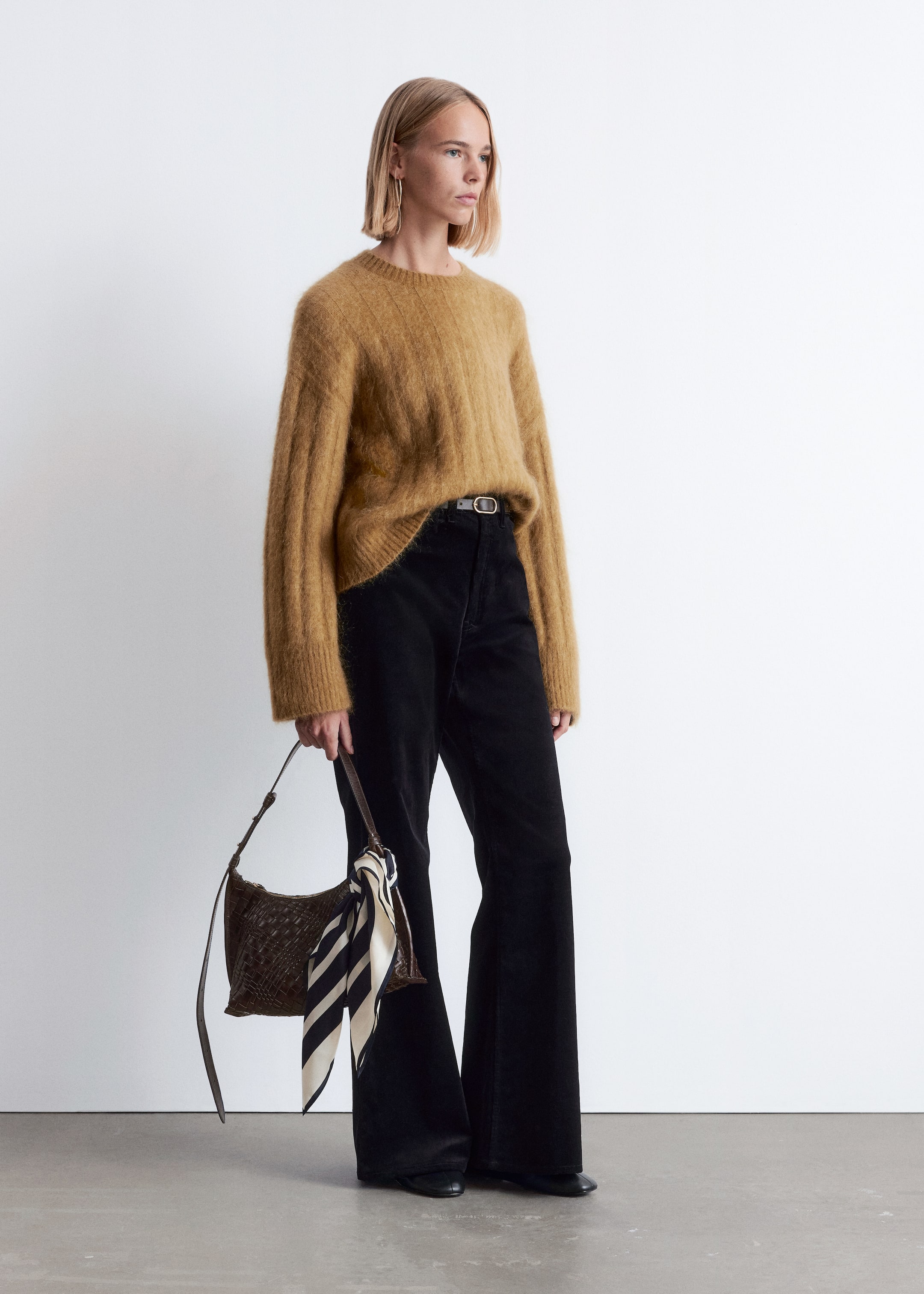 Größeres Bild anzeigen: Eine Ganzkörperansicht zeigt ein Outfit, das einen hellbraunen, flauschigen Pullover mit Rundhalsausschnitt und subtiler vertikaler Rippung sowie einer lockeren Passform präsentiert, kombiniert mit schwarzen, weit geschnittenen Cordhosen. Ein dünner grauer Gürtel betont die Taille, und eine dunkelbraune geflochtene Tasche mit einem gestreiften Schal wird gehalten, das Ensemble mit schwarzen flachen Schuhen vervollständigend.