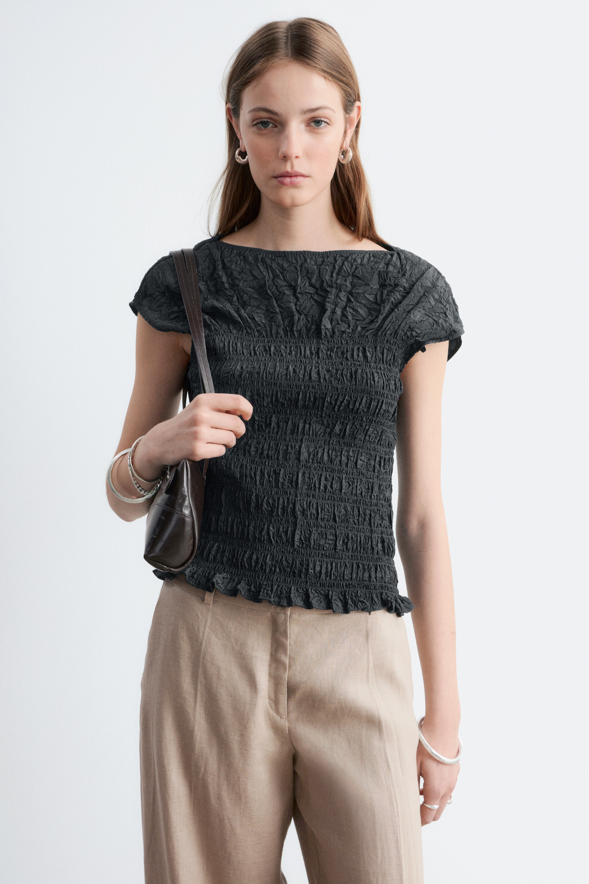 Agrandir l'image: Le haut en charbon foncé présente un large décolleté bateau et des manches courtes à capuchon. Son corps est entièrement froncé, créant un aspect ajusté et texturé, avec un tissu froissé sur le haut de la poitrine et les épaules, et un ourlet subtilement volanté.