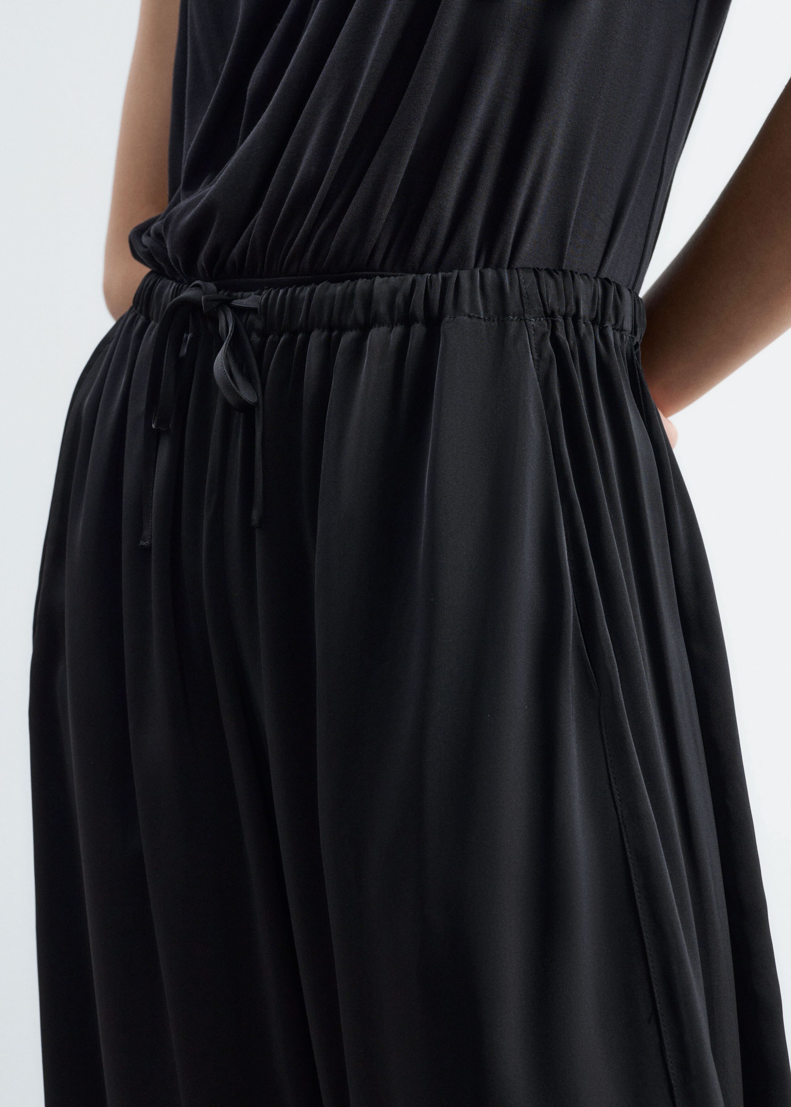 The model is wearing black fluid trousers featuring a gathered elasticated waistband with a drawstring tie. The loose-fitting trousers are styled with a sleeveless black top tucked into the waistband.