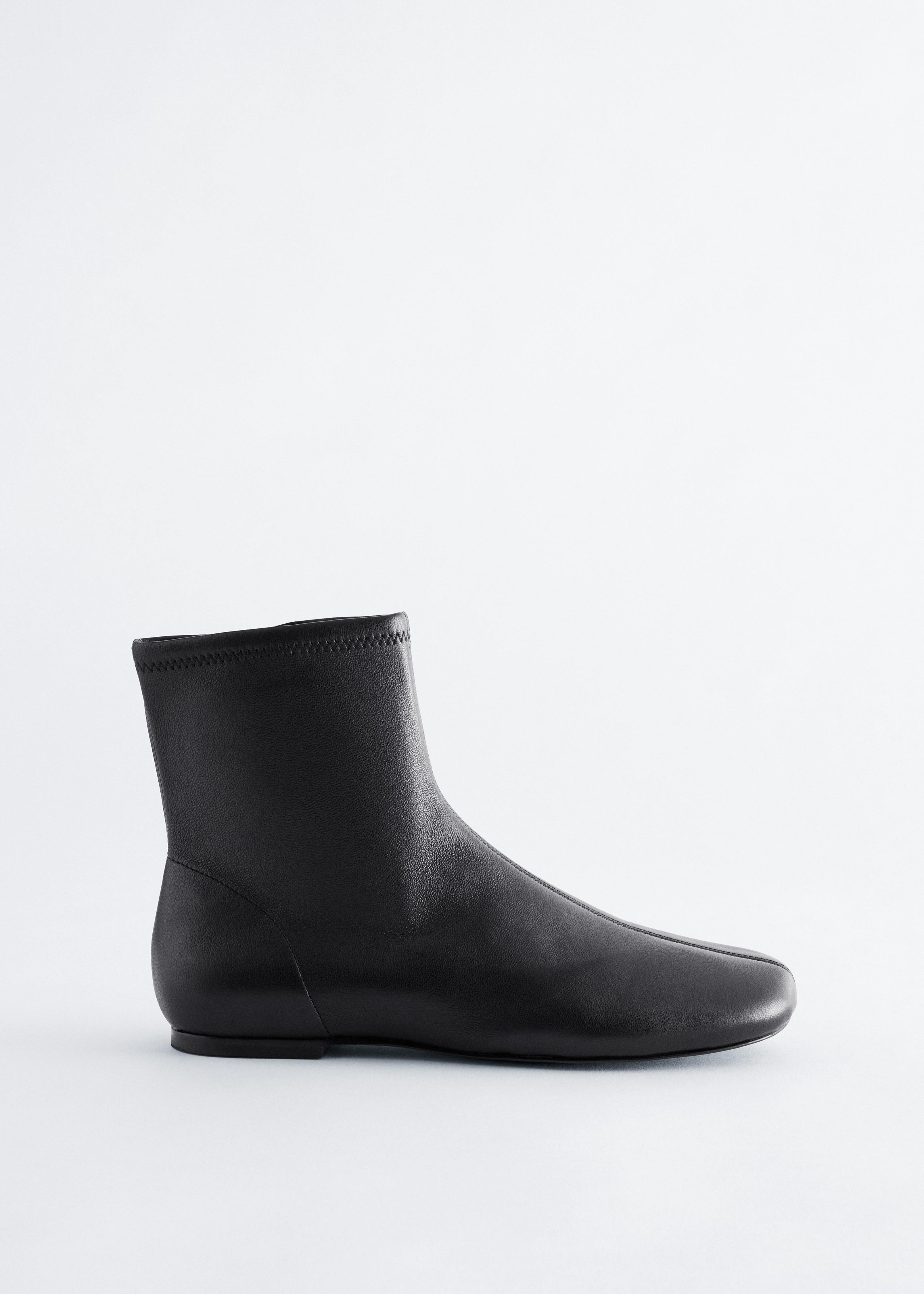 Black leather boots featuring a rounded toe and central seam are worn by the model, with wide-leg black trousers draping over them.