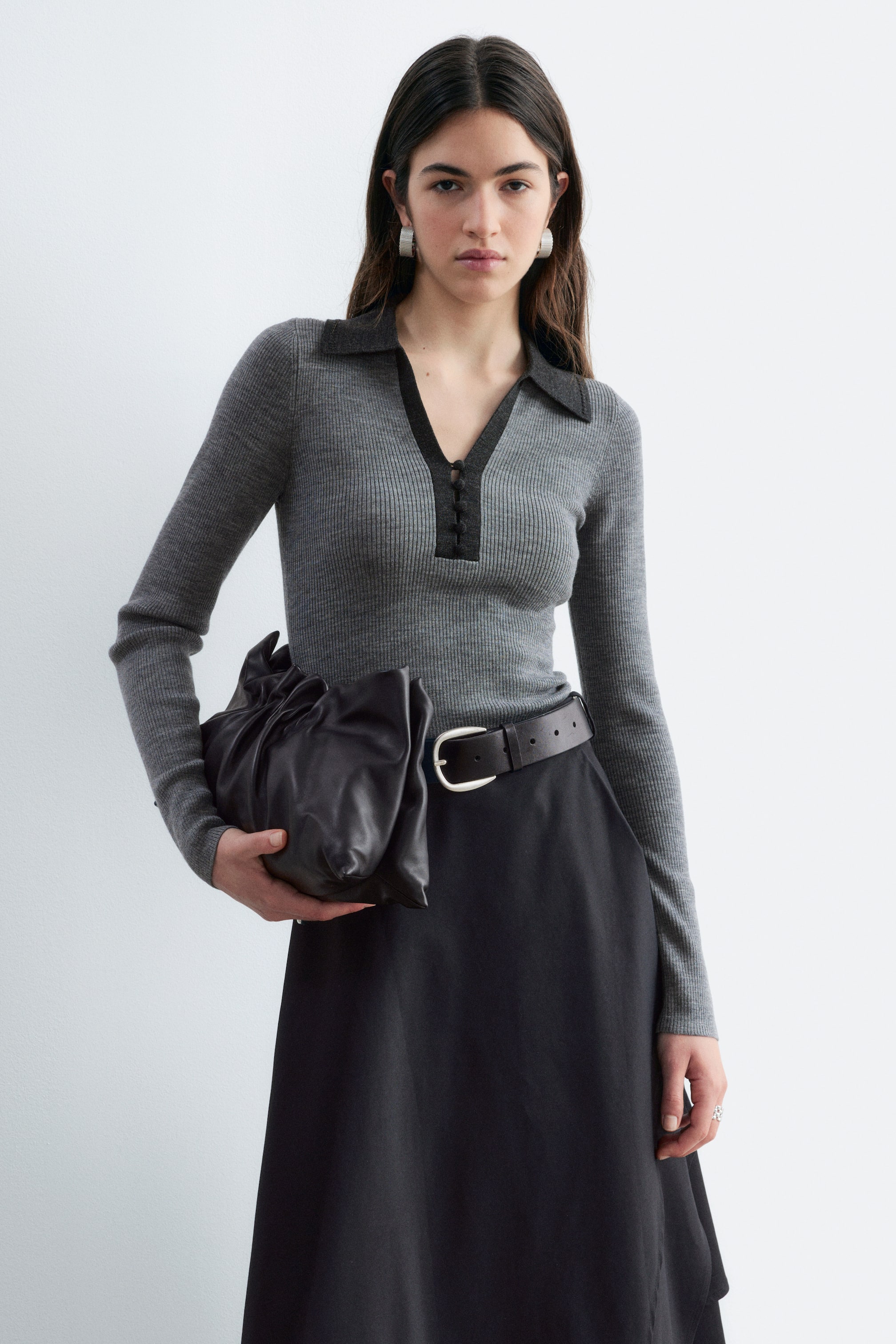 Visualizza l'immagine ingrandita: Un top in lana grigia a coste, dalla vestibilità aderente e con maniche lunghe, indossato dalla modella, caratterizzato da un colletto in contrasto grigio scuro e da uno scollo a V aperto con bottoni rivestiti in tessuto. Il top è infilato in una gonna nera con cintura in pelle scura, mentre la modella tiene in mano una clutch nera in pelle arricciata.