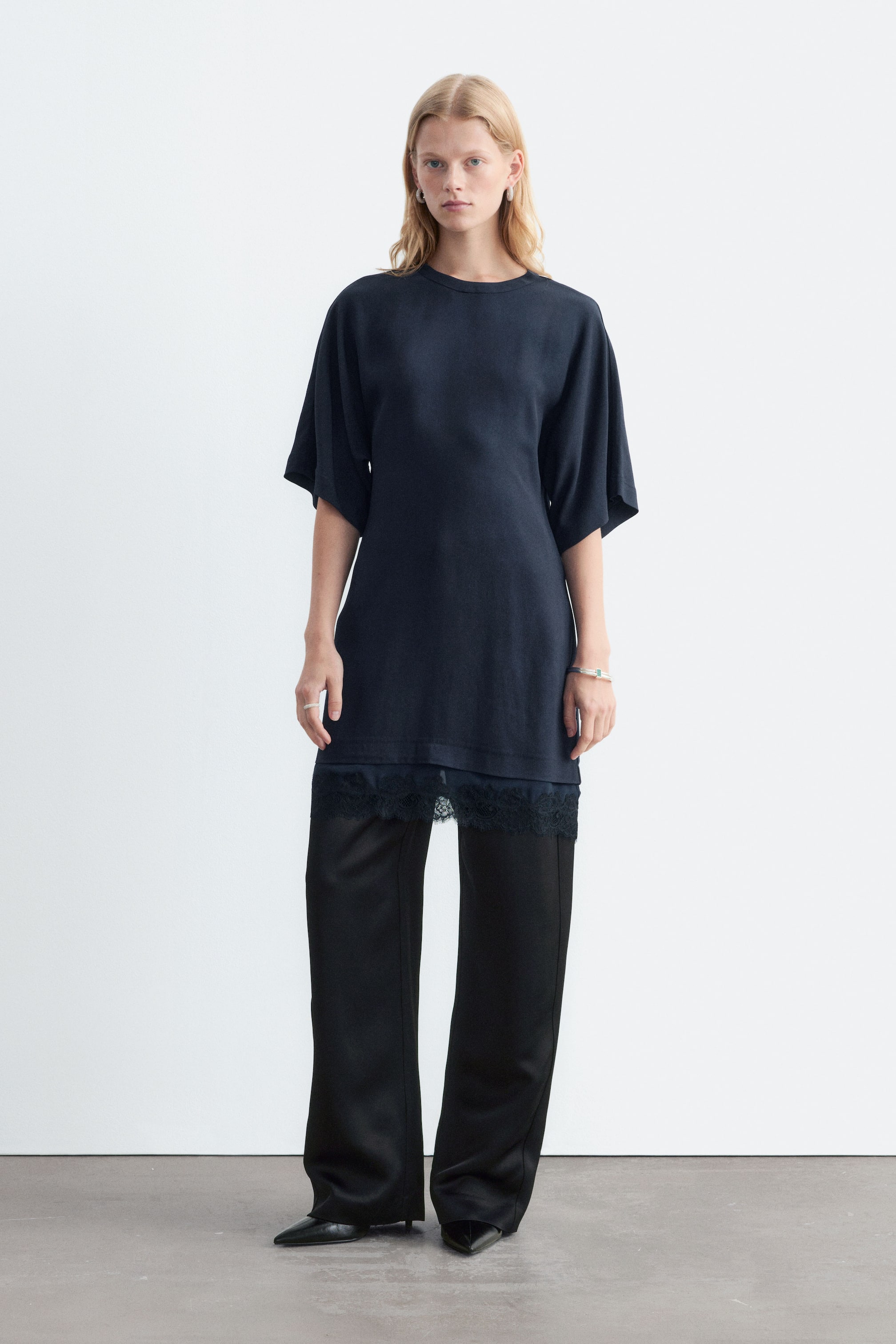 View larger image: The model is wearing a navy blue short-sleeved dress with a round neckline and a delicate lace panel at the hem. The dress is styled as a tunic layered over black wide-leg trousers.