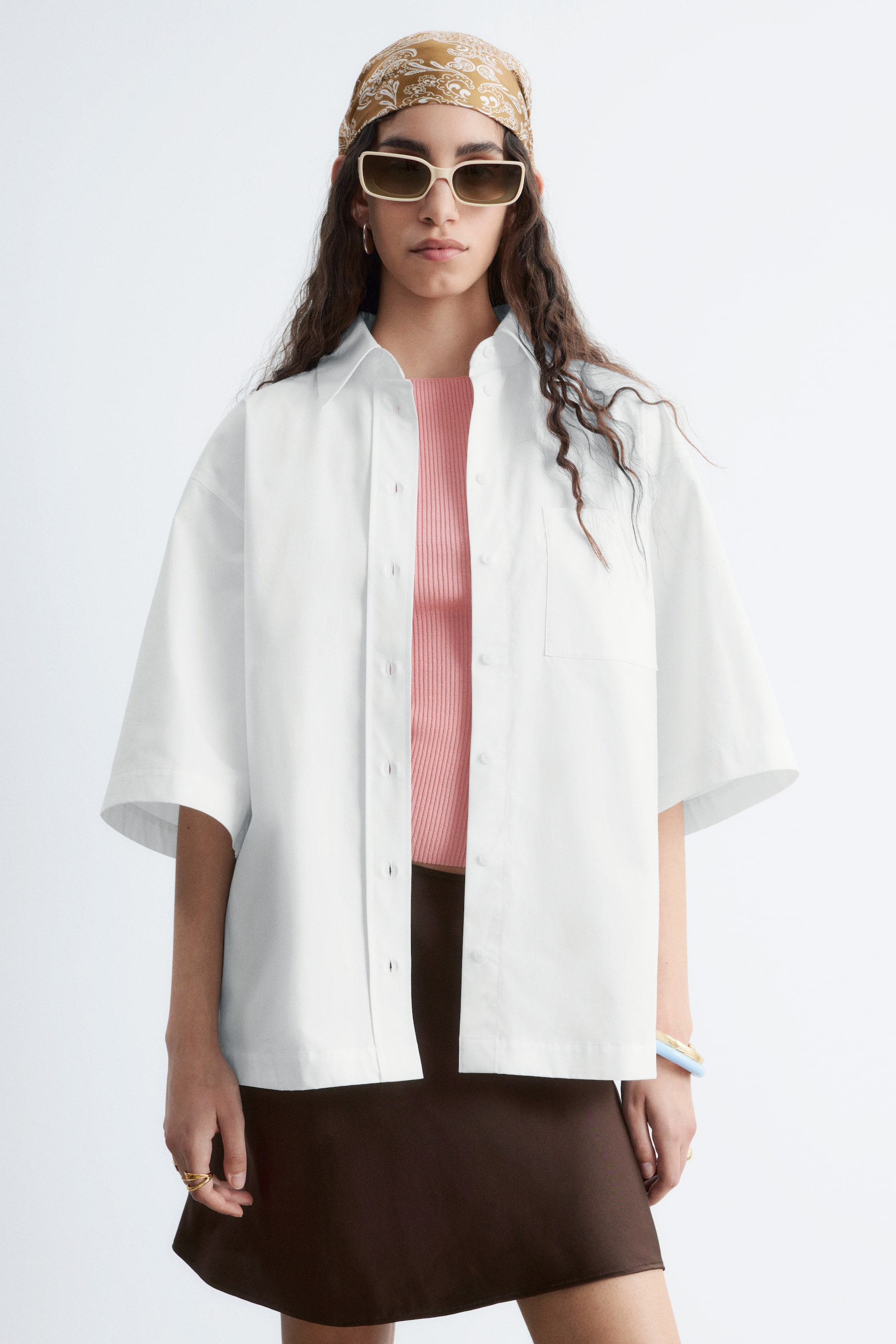 Προβολή μεγαλύτερης εικόνας: Η μοντέλο φοράει ένα λευκό πουκάμισο με κοντά μανίκια. Ο γιακάς του πλαισιώνει το ντεκολτέ, ενώ το μπροστινό μέρος του είναι μερικώς ξεκουμπωμένο και μια μονή τσέπη στο στήθος βρίσκεται στην αριστερή πλευρά. Το πουκάμισο είναι στυλιζαρισμένο ανοιχτό πάνω από ένα ροζ ριμπ τοπ.