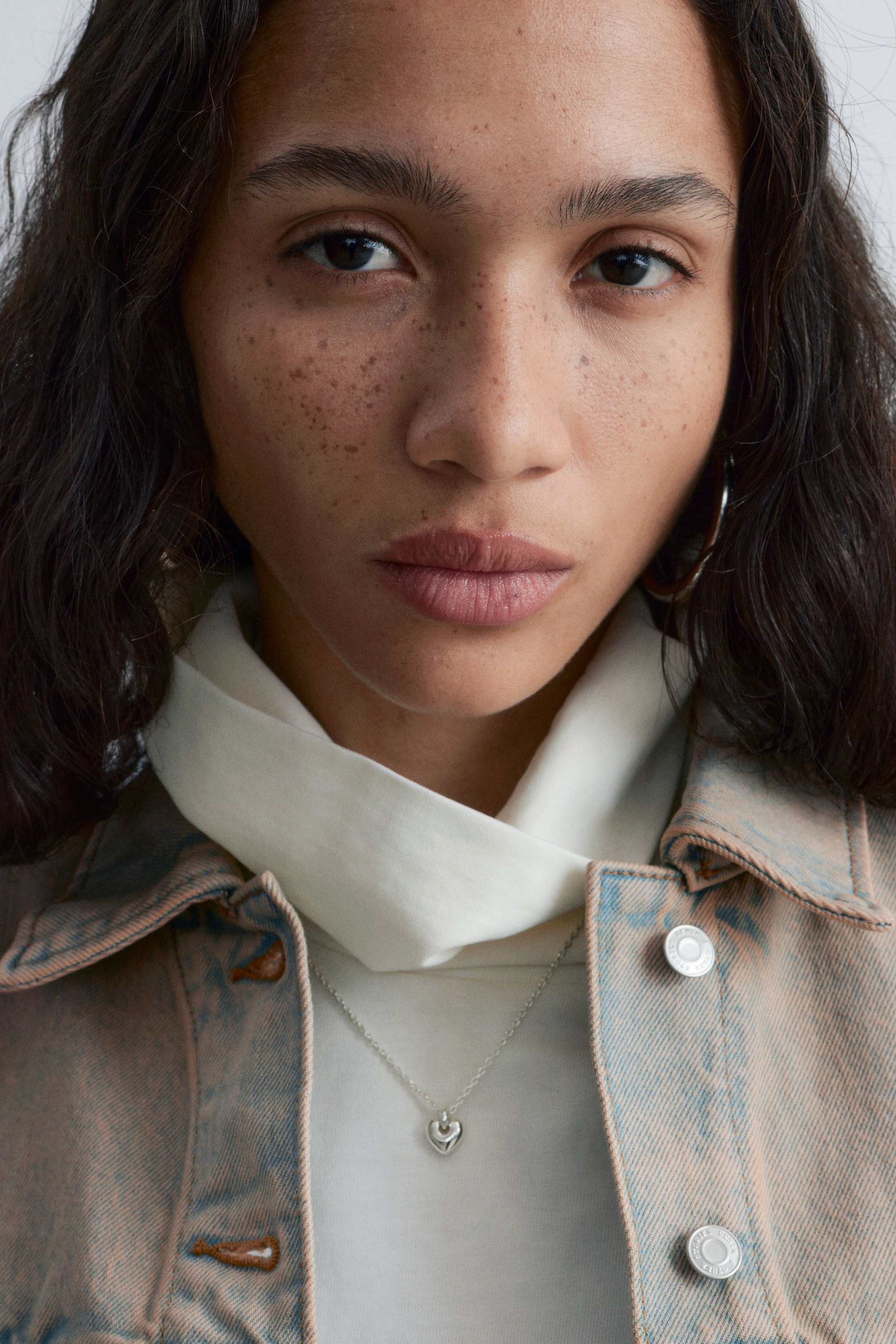 Ver imagen más grande: Una chaqueta vaquera de lavado ácido, con un tono azul claro y rosado, se lleva abierta sobre un top de cuello alto color crema. Un collar de cadena plateada con un pequeño colgante en forma de corazón descansa centralmente sobre el top crema.