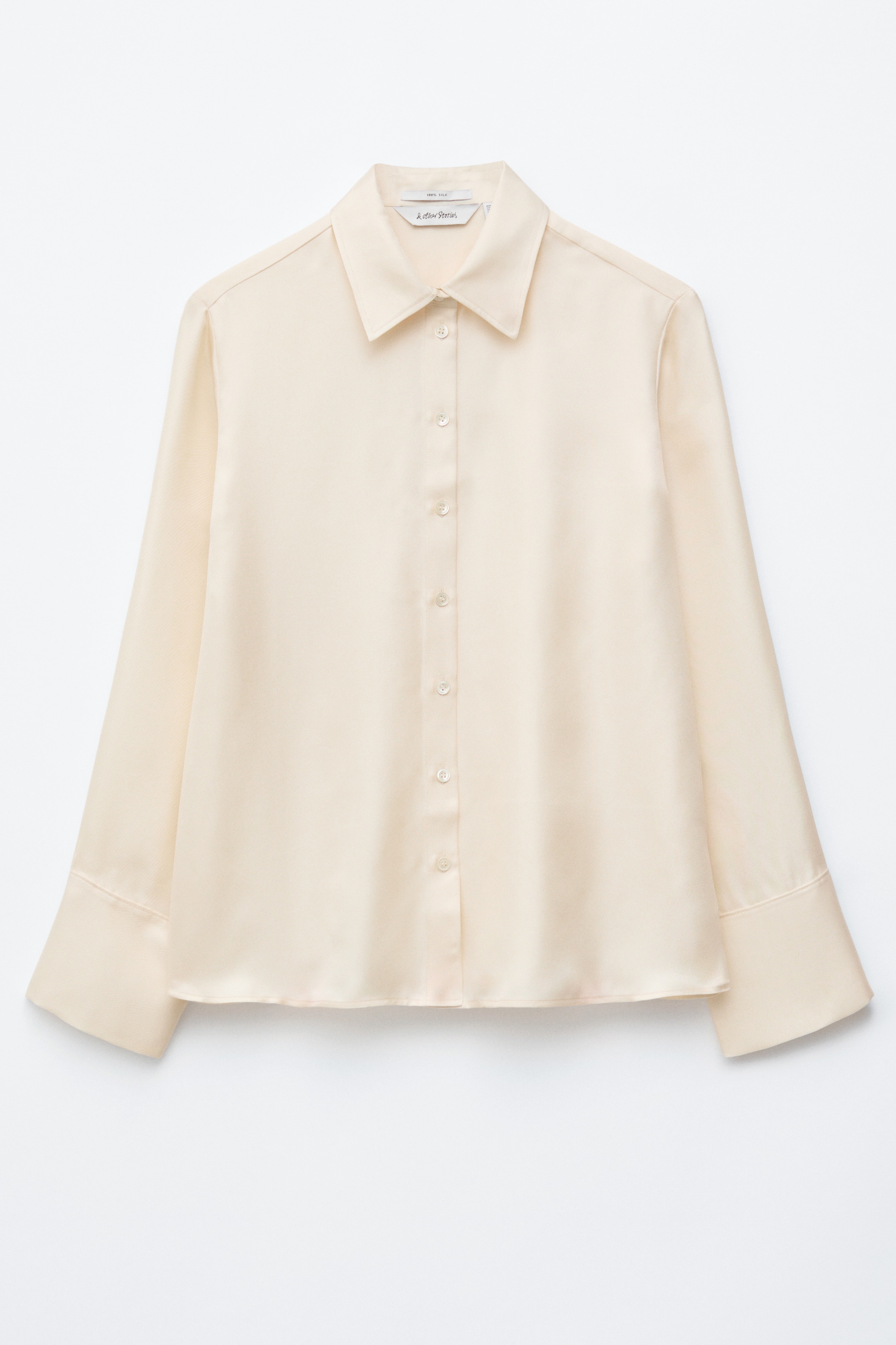 Ver imagen más grande: Una camisa de seda color crema presenta su PARTE DELANTERA, con un acabado lustroso y una silueta relajada. Está diseñada con un cuello clásico, una parte frontal abotonada y puños anchos, cada uno asegurado por un solo botón.