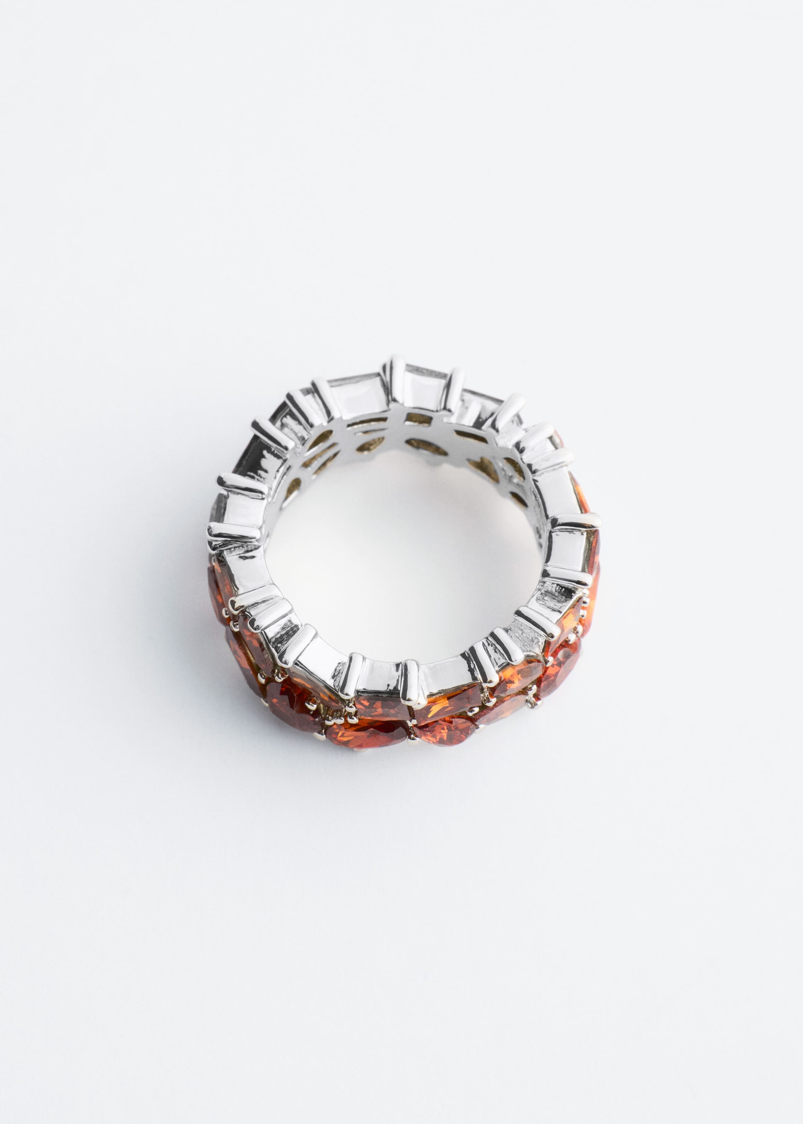 A silver-tone band ring is resting at an angle, displaying a double row of faceted, oval-cut orange gemstones secured in prong settings. The top-down view shows the structured, polished interior of the band with open metalwork behind the gemstones.