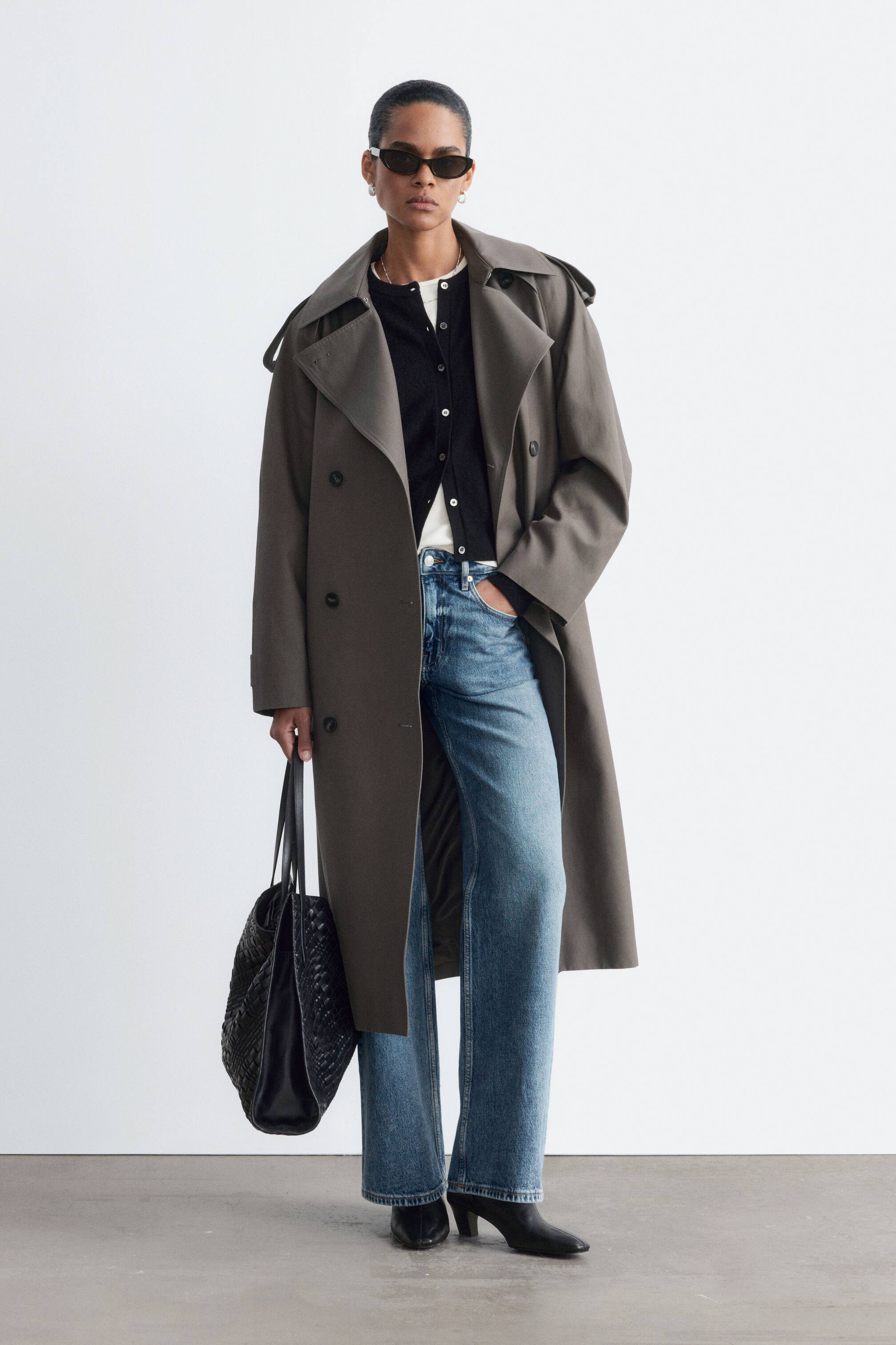Größeres Bild anzeigen: Ein locker geschnittener, zweireihiger Trenchcoat in dunklem Taupe wird vom Model offen getragen und betont die breiten Revers, die Schulterklappen und die Ärmellaschen. Der Mantel wird über einem schwarzen Cardigan mit Knopfleiste, einem weißen T-Shirt und einer blauen Jeans gestylt, während das Model eine schwarze, gewebte Tote Bag trägt.
