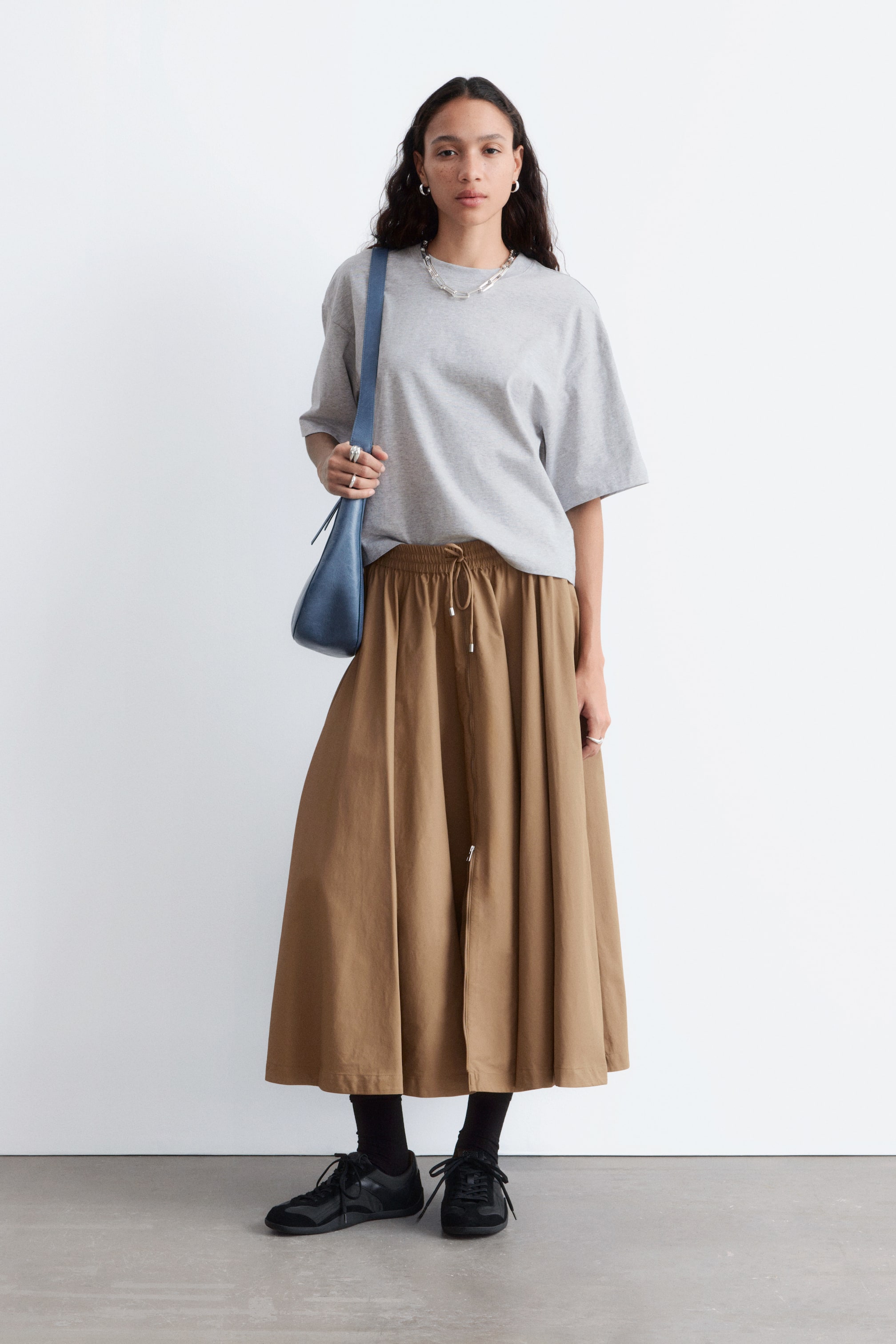 Agrandir l'image: Une jupe midi marron en forme de A avec des plis et une taille à cordon est portée par le mannequin, qui rentre un t-shirt gris dans sa taille.