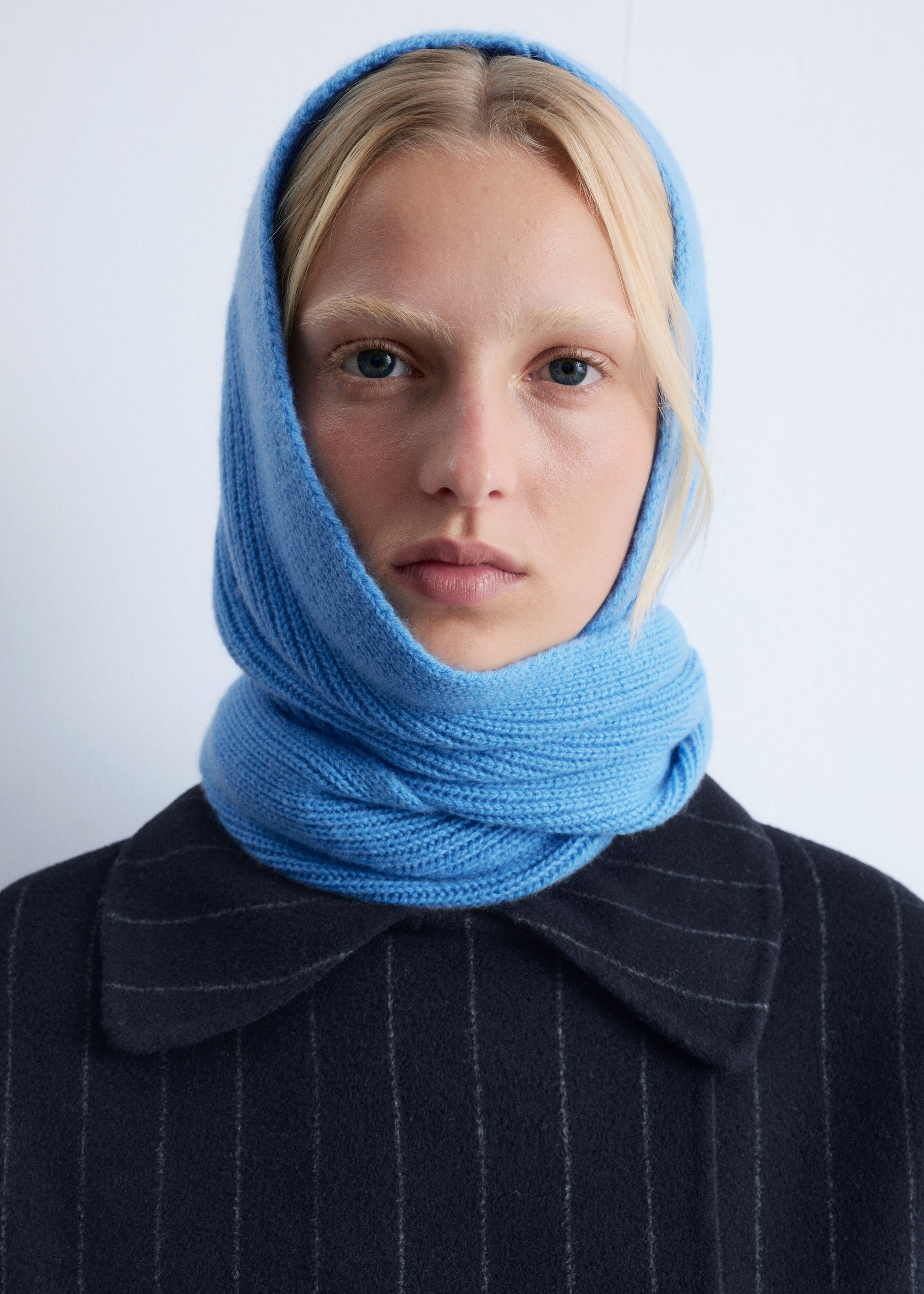 Visualizza immagine più grande: Uno snood a maglia di un blu brillante viene indossato, caratterizzato da una texture a coste. Copre la testa e le orecchie, attorcigliandosi intorno al collo, abbinato a una giacca scura a righe sottili.
