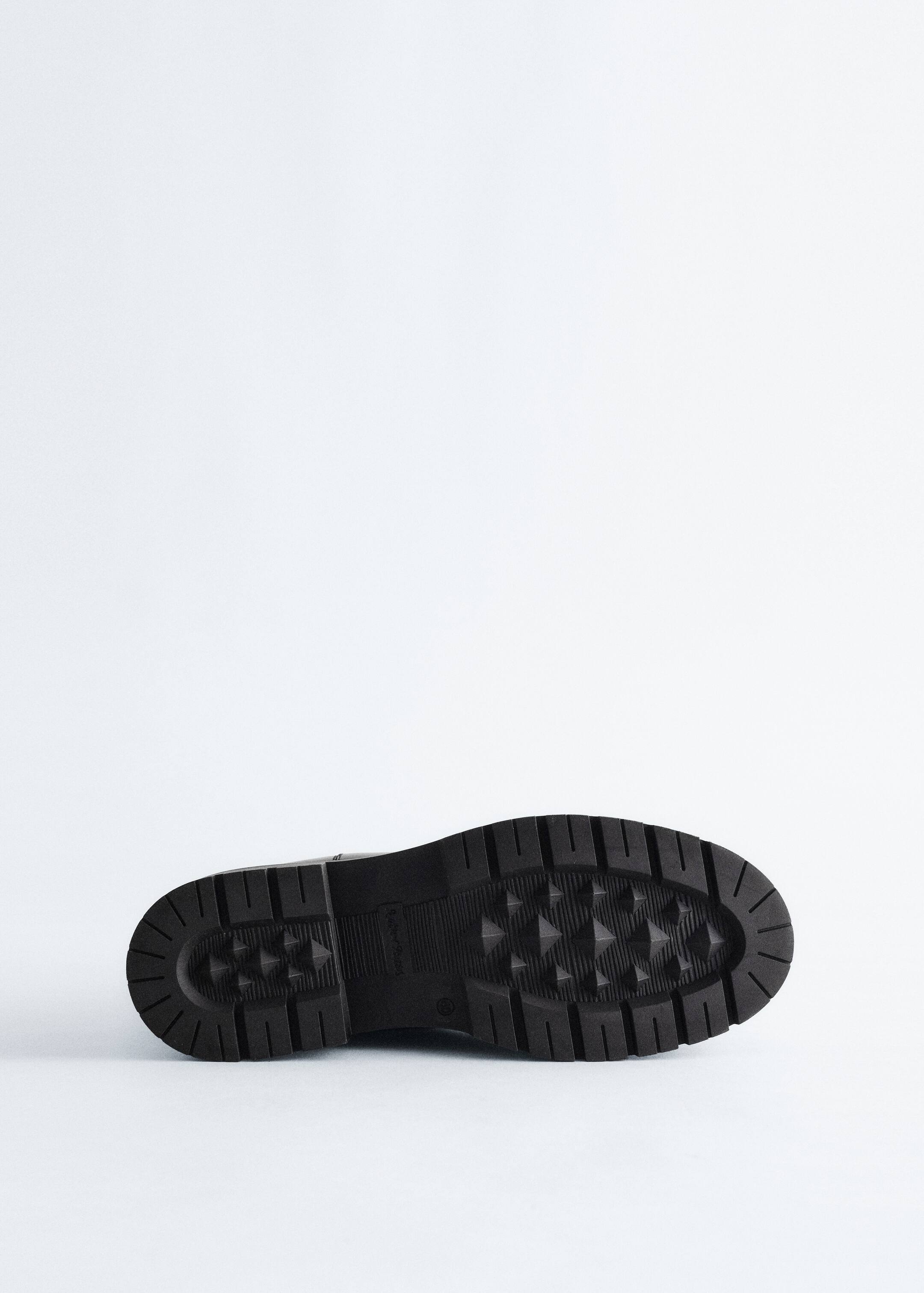 Grösseres Bild ansehen: Ein schwarzer Stiefel wird von UNTEN betrachtet und zeigt seine klobige, gemusterte Sohle. Die Sohle weist tiefe rechteckige Stollen entlang des Randes und pyramidenförmige Nieten im zentralen Bereich auf.