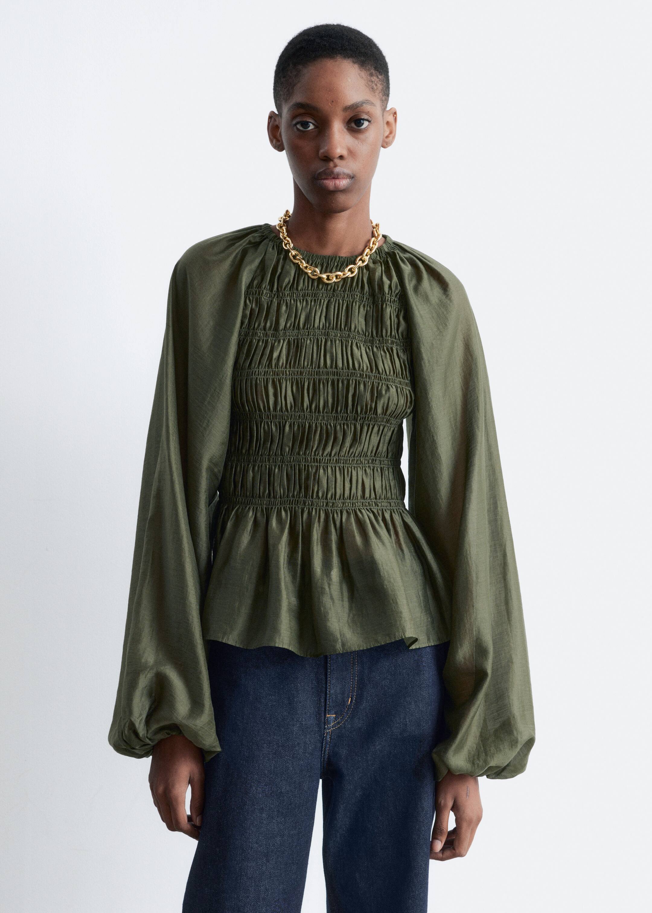 View larger image: A lustrous olive green blouse is being worn by the model. It features a smocked body that flares into a peplum hem, a round gathered neckline, and raglan balloon sleeves with elasticated cuffs, and is styled with dark blue jeans and a gold-colored chain necklace.