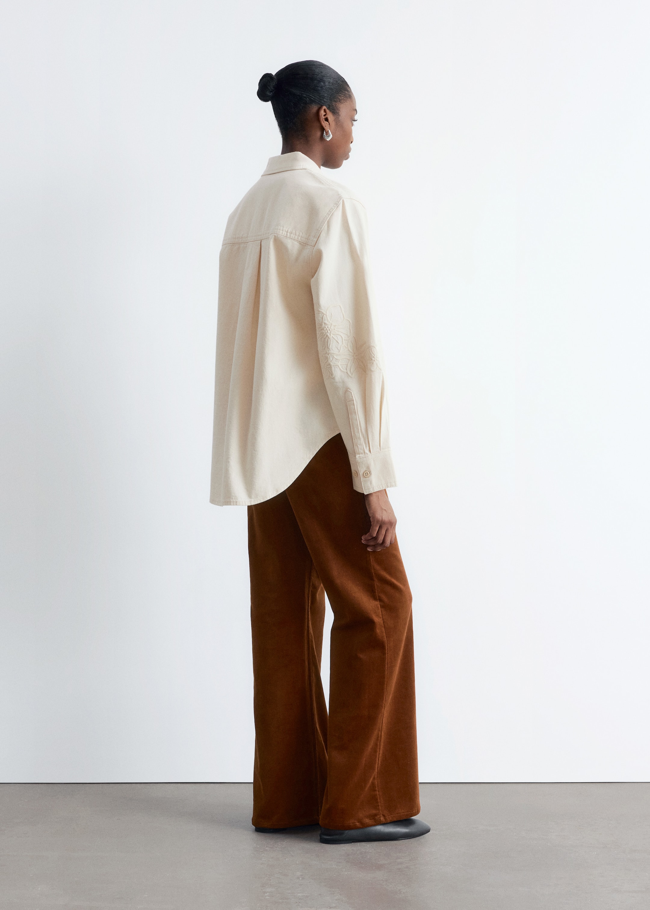 Zobrazit větší obrázek: Krémová džínová košile s vyšívanými ozdobami na horním rukávu je nošena modelem. Volně padnoucí košile má klasický límec a dlouhé rukávy a je stylizována s hnědými kalhotami s širokými nohavicemi.