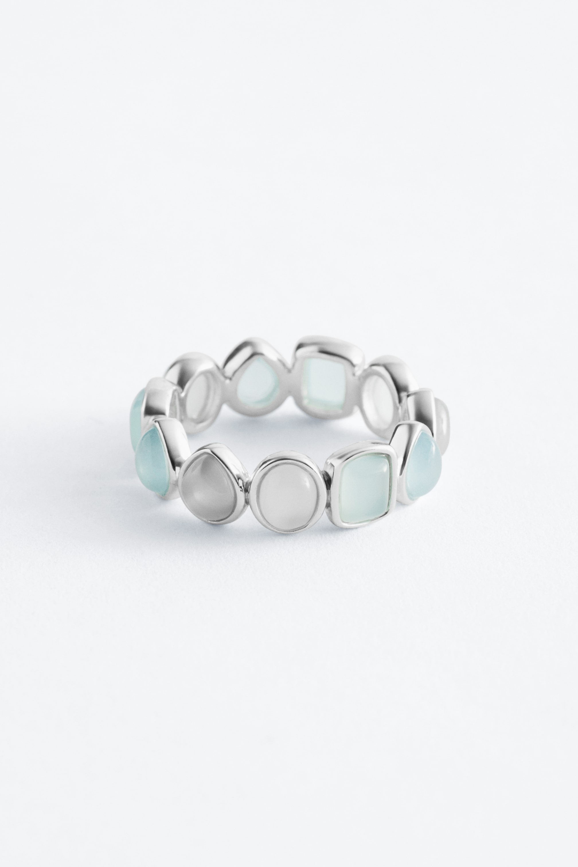 Visualizza immagine più grande: Un anello dal tono argento presenta una fascia continua di pietre di varie tonalità di azzurro chiaro e bianco latteo. Queste pietre appaiono in forme assortite, inclusi cerchi, quadrati e gocce, e sono disposte strettamente attorno alla circonferenza dell'anello.