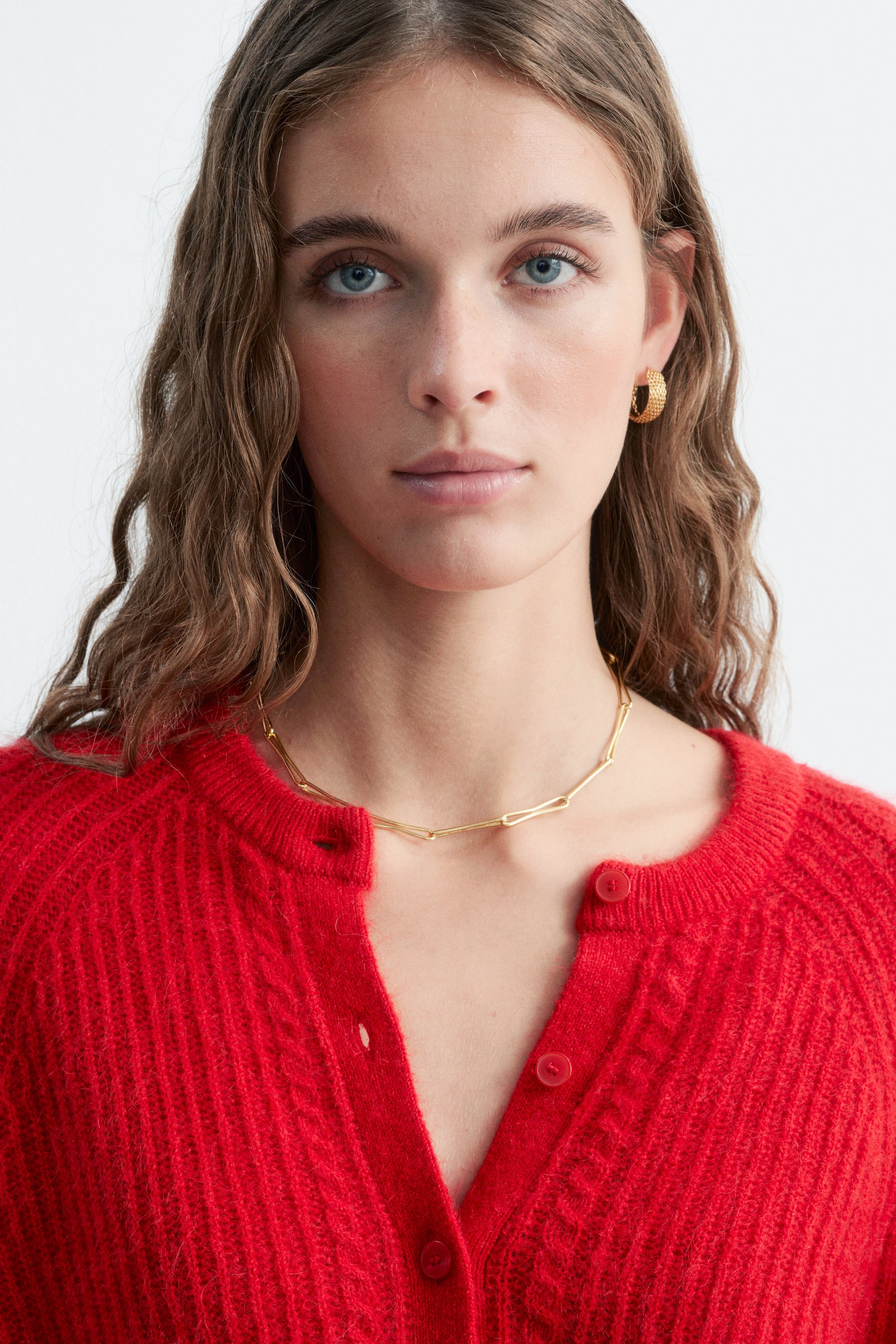 Visualizza immagine più grande: La modella indossa un cardigan rosso testurizzato con lavorazione a maglia a coste e dettagli a trecce lungo l’abbottonatura. Il cardigan è indossato con il collo parzialmente sbottonato, mettendo in risalto una sottile collana a catena dorata.