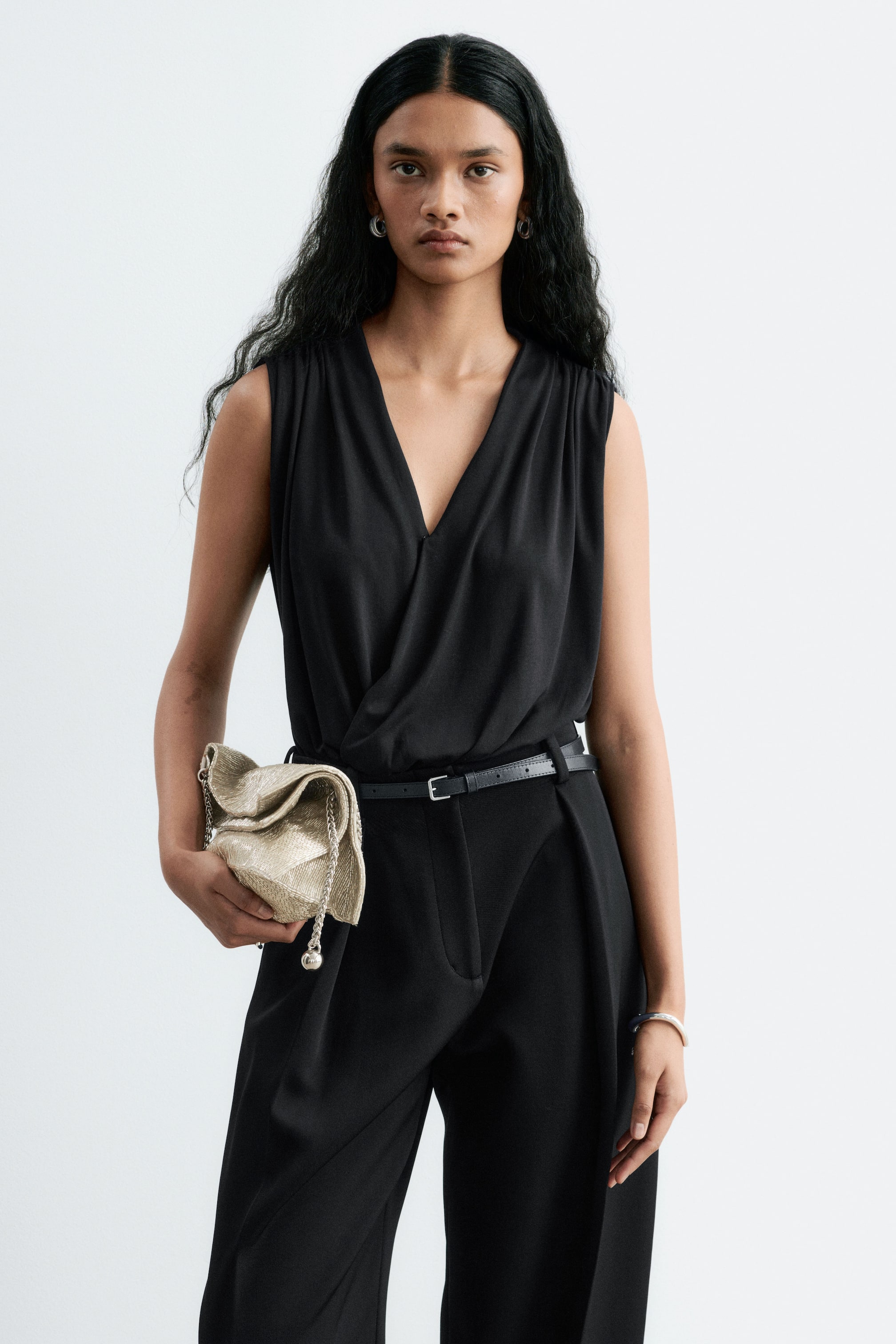 Grotere afbeelding bekijken: Een zwarte mouwloze bodysuit met een gedrapeerde overslag-V-hals en geplooide schouders wordt gedragen door het model en is ingestopt in een zwarte broek met hoge taille en een dunne zwarte riem. Het model houdt een gestructureerde metallic gouden clutch aan de heup vast.
